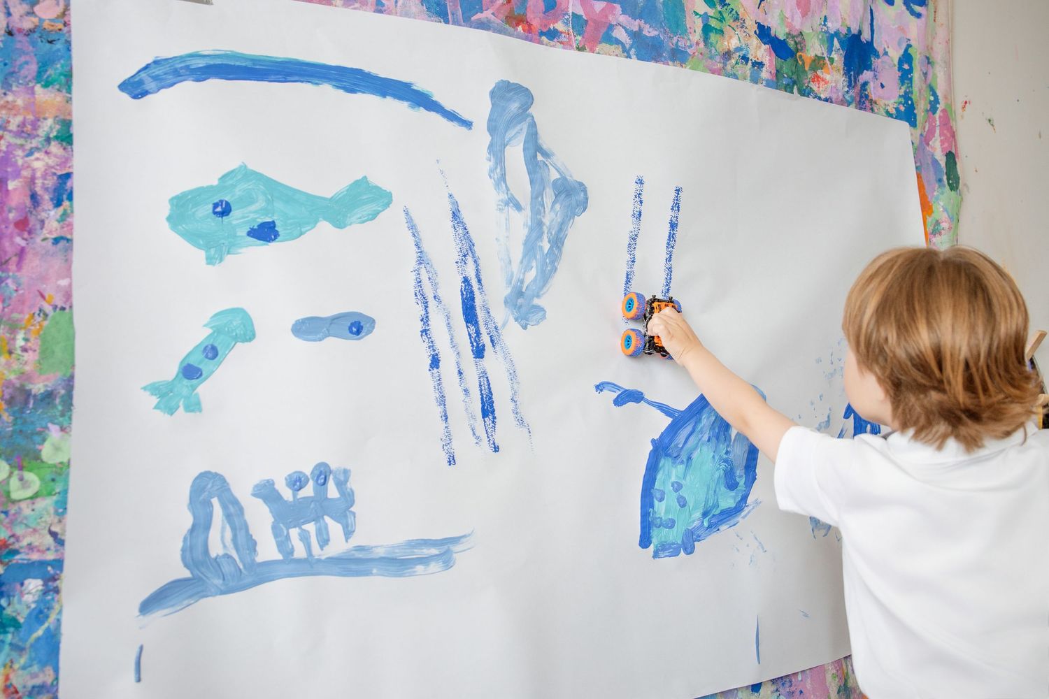 Young child painting blue abstract shapes on large white canvas with colorful painted backdrop.