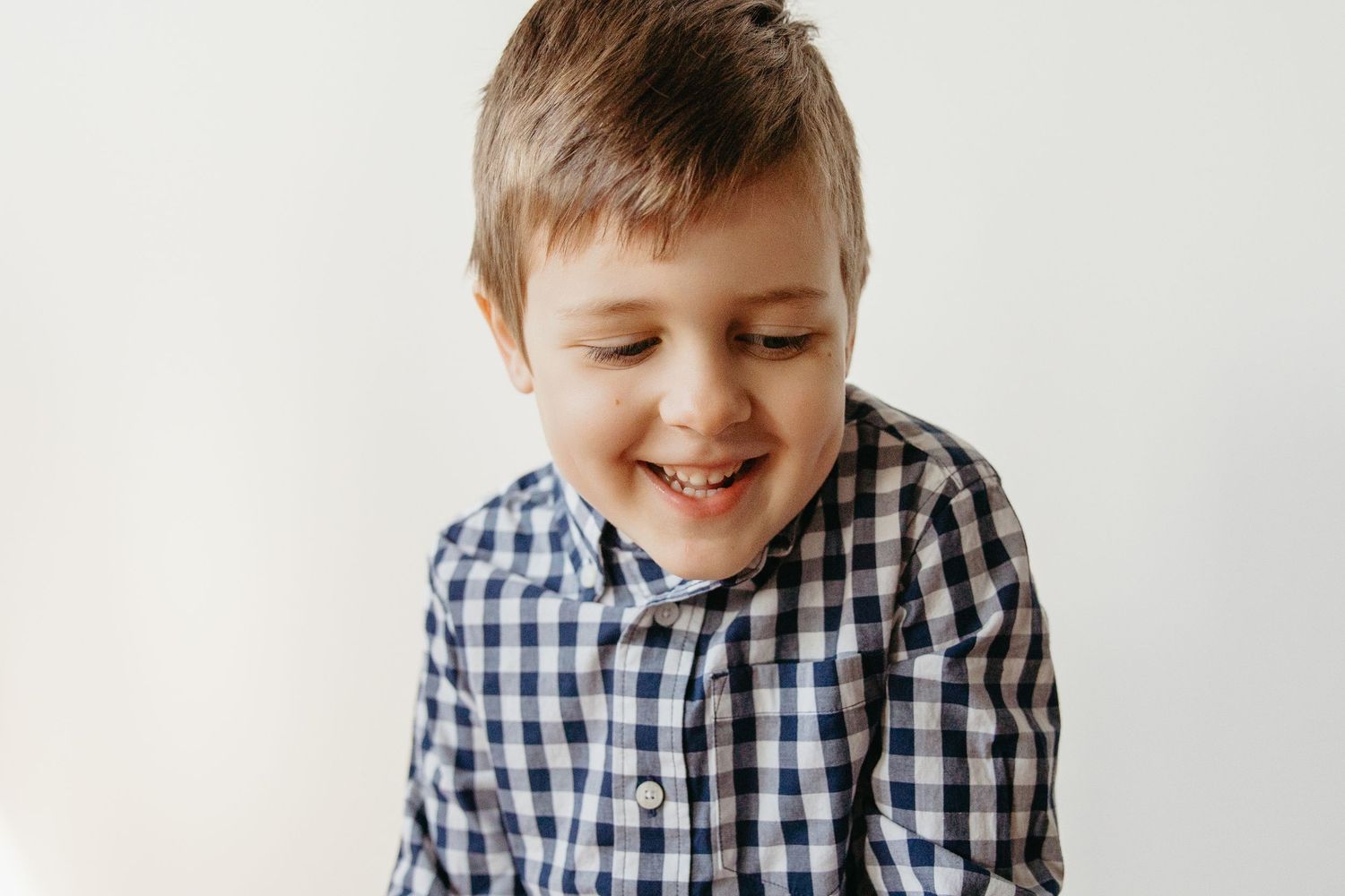 A cheerful young boy in a blue plaid shirt laughs with his eyes closed against a white background.