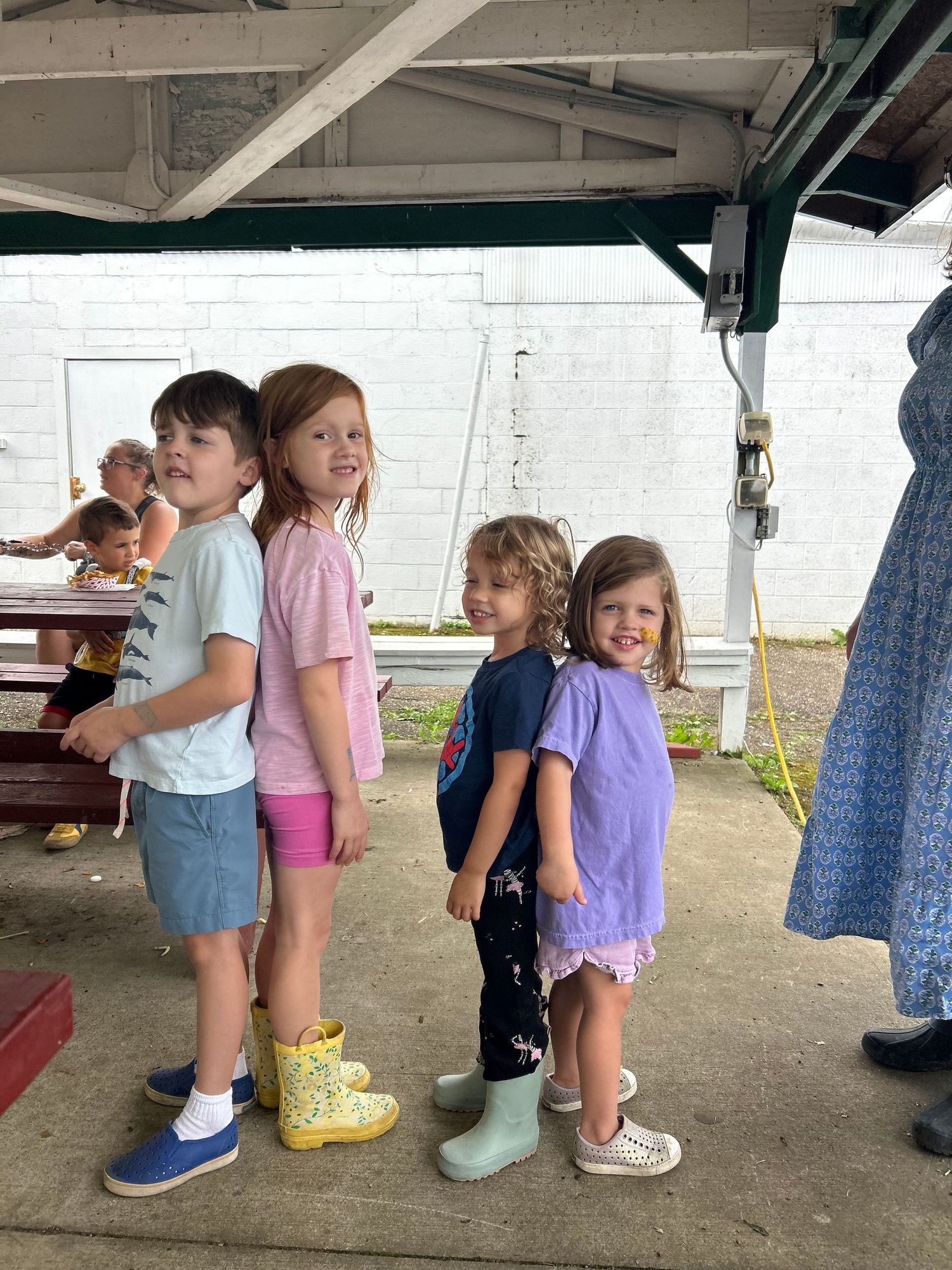 Four young children standing in a line under a covered outdoor area, wearing colorful boots and casual summer clothing.