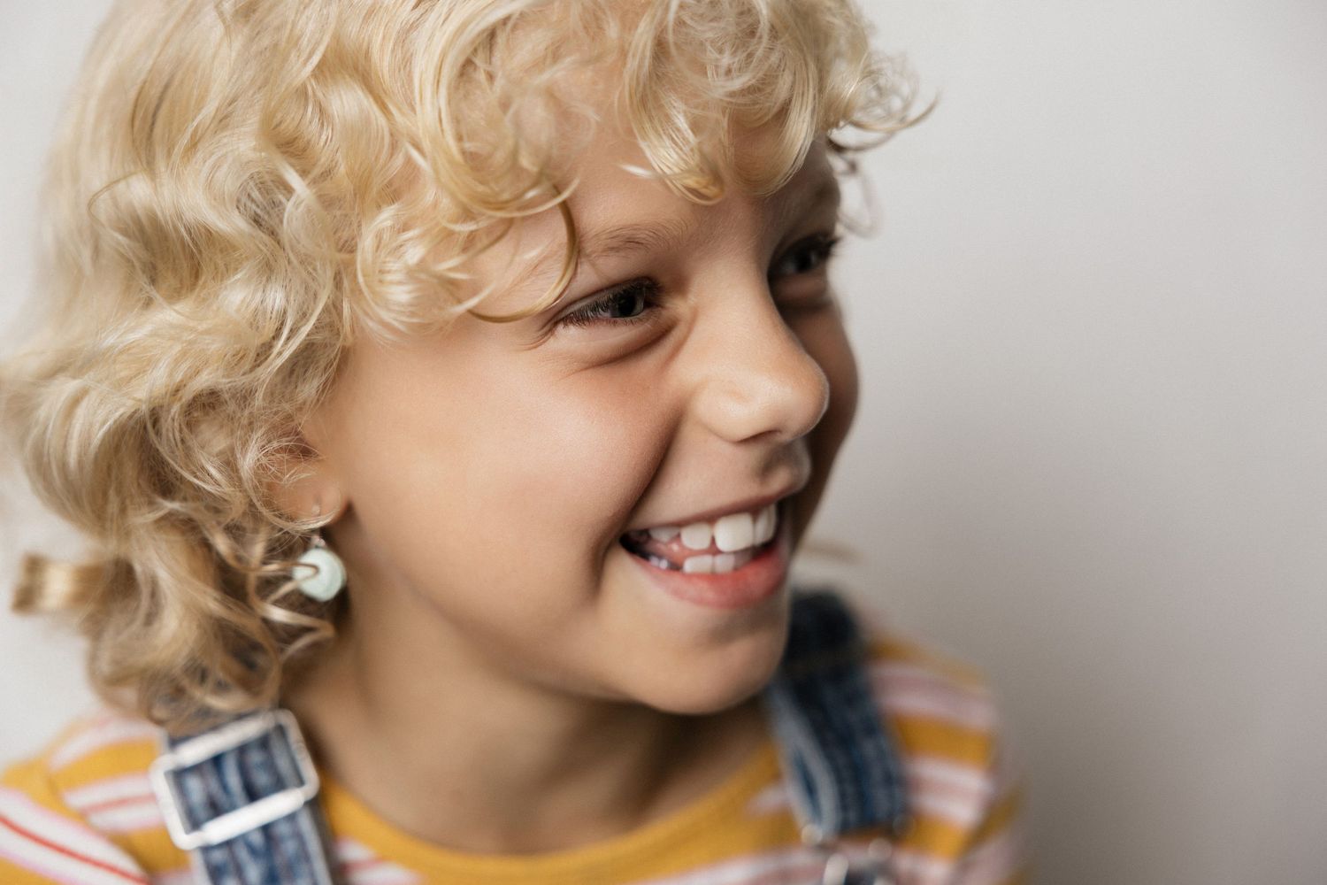 Smiling blond curly-haired child wearing striped shirt and denim overalls, laughing joyfully against white background.