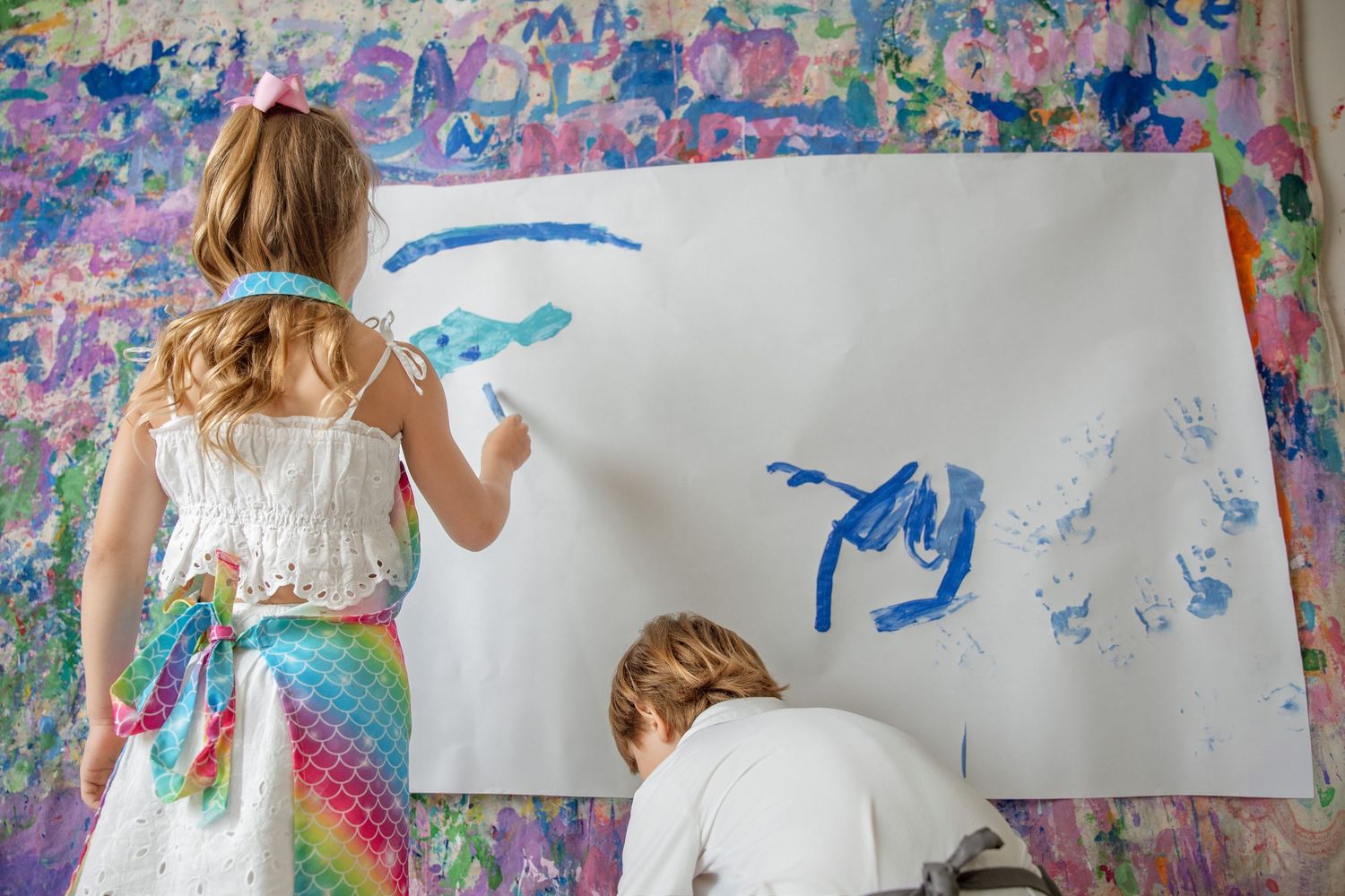 Two children painting blue designs on white paper against a colorful painted wall backdrop.