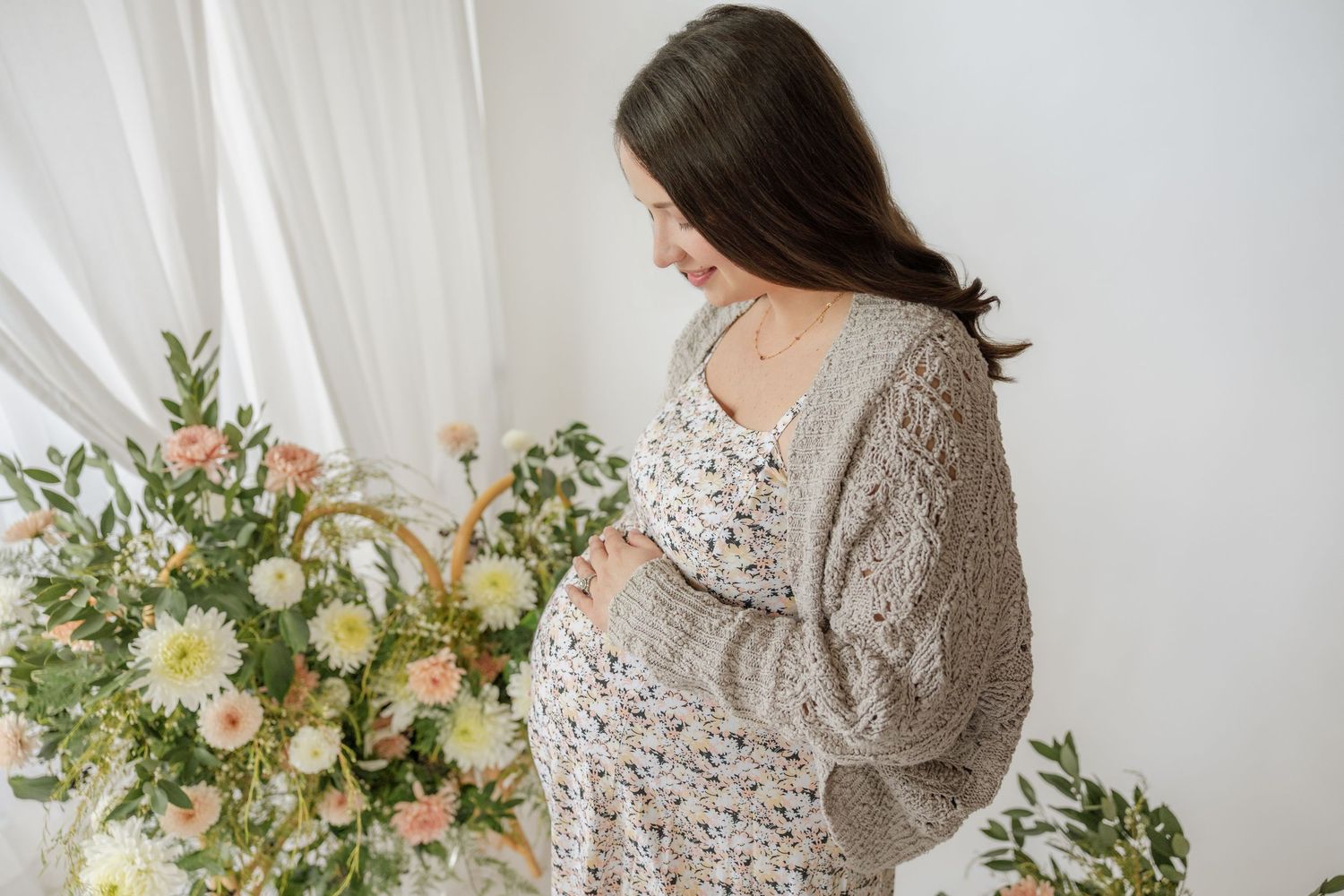 A pregnant woman in a floral dress and cardigan cradles her baby bump near a window with flowers.