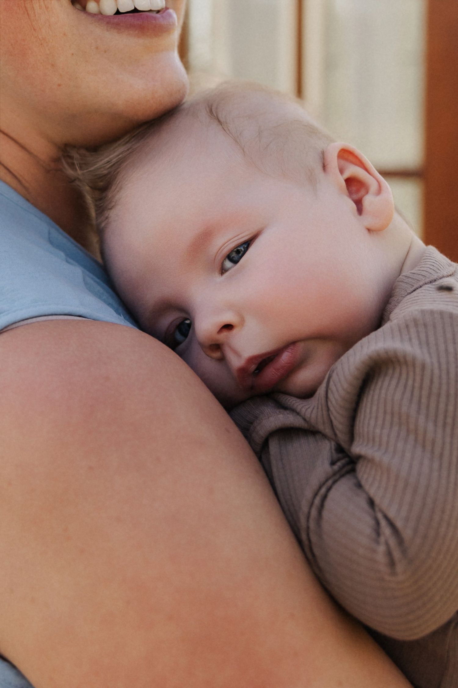 A smiling mother tenderly holds her sleepy newborn baby against her chest, the infant gazing softly in a cozy brown outfit.