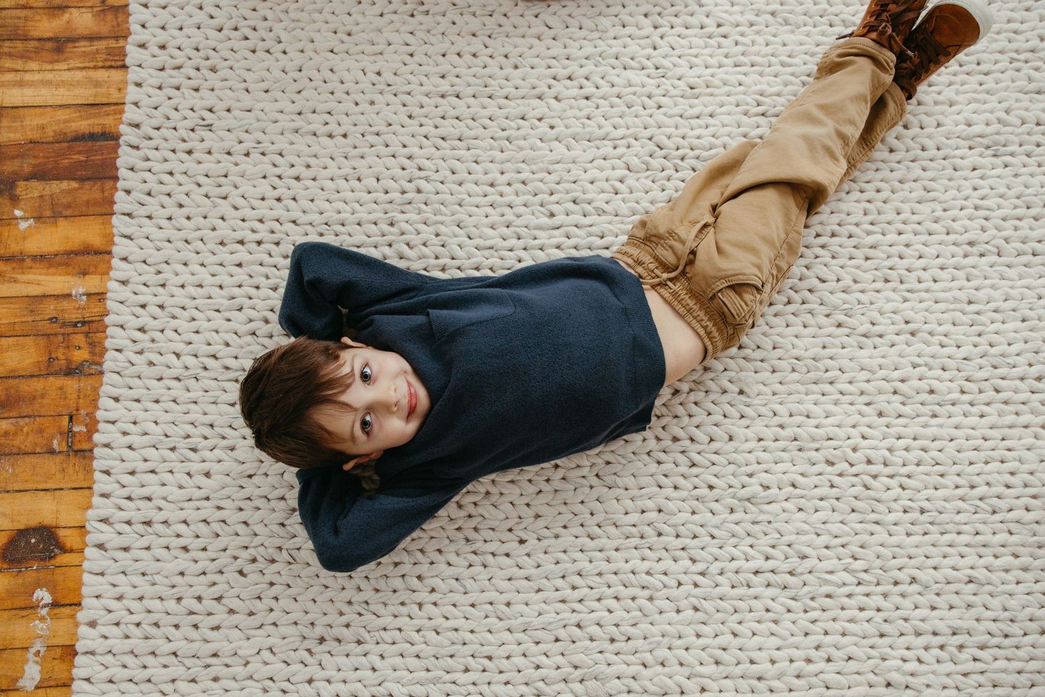Young boy lying on a chunky knit rug viewed from above wearing navy sweater.