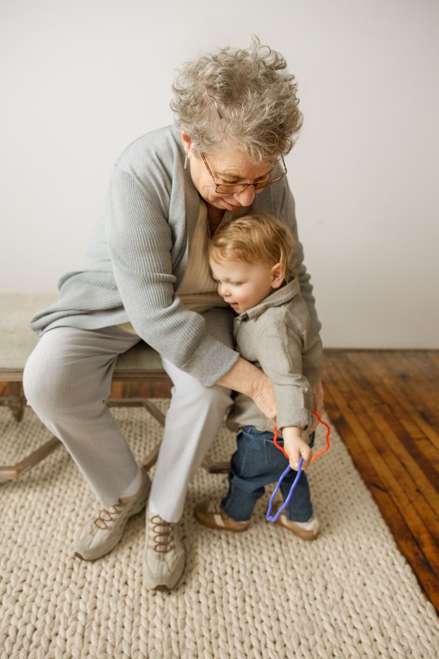Elderly grandmother sitting with toddler on her lap, helping child play with colorful toy on textured rug.