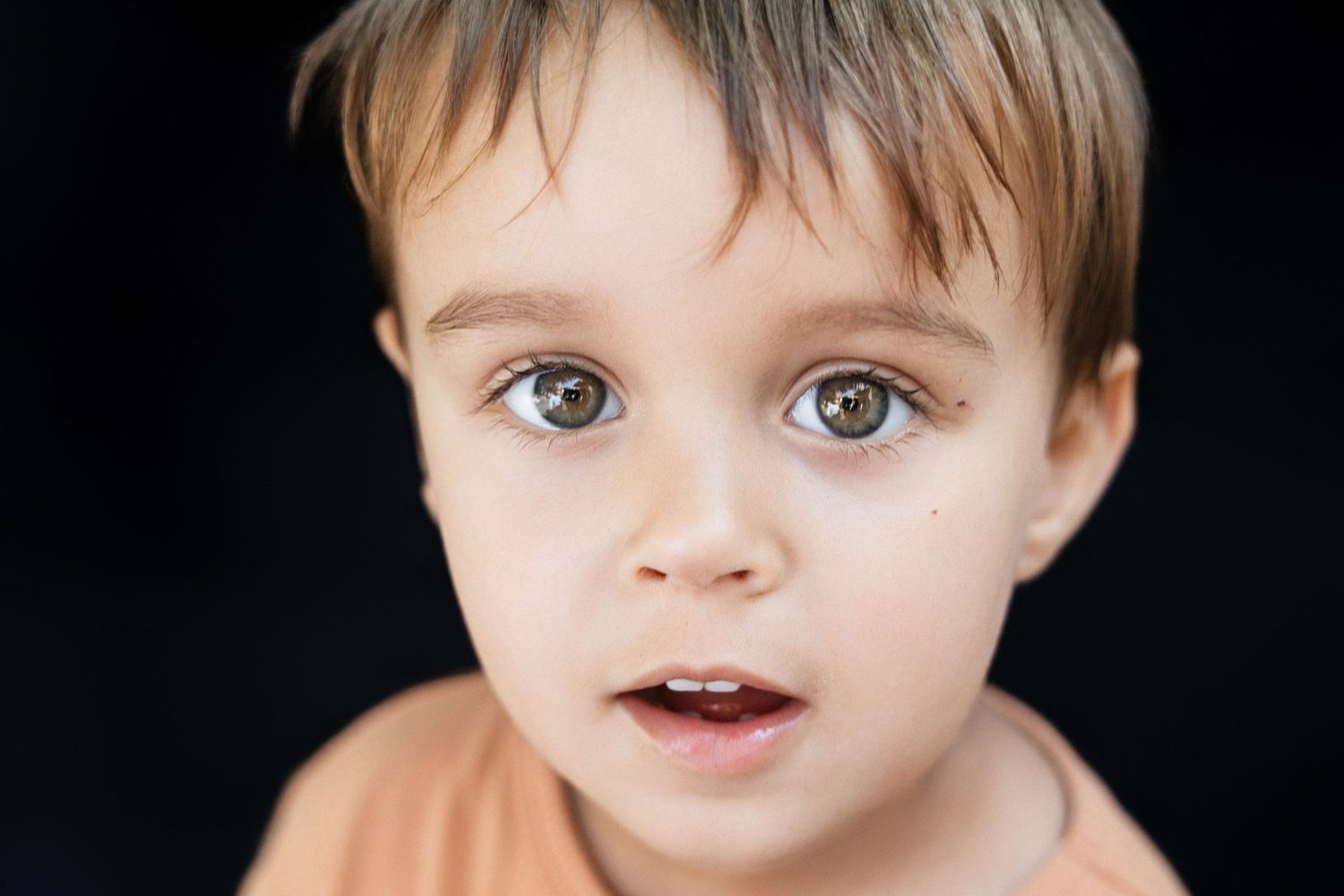 Close-up portrait of a young toddler boy with big brown eyes and light brown hair on dark background.