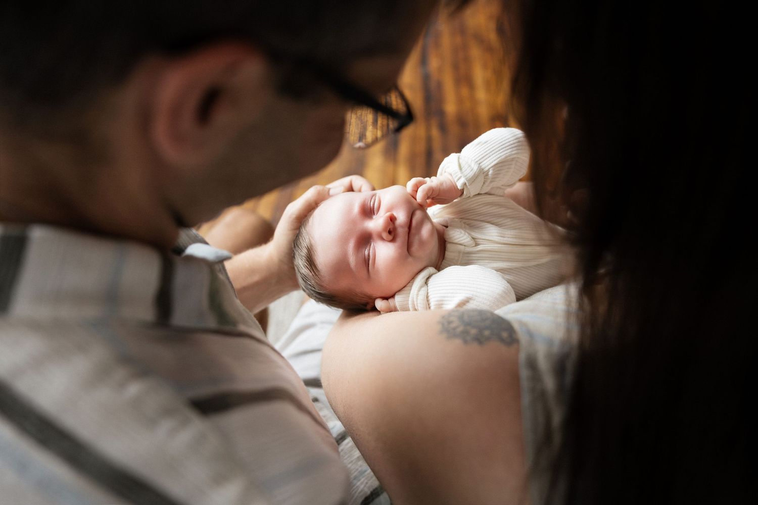 Parents lovingly gazing at their sleeping newborn baby lying on a soft surface in warm light.