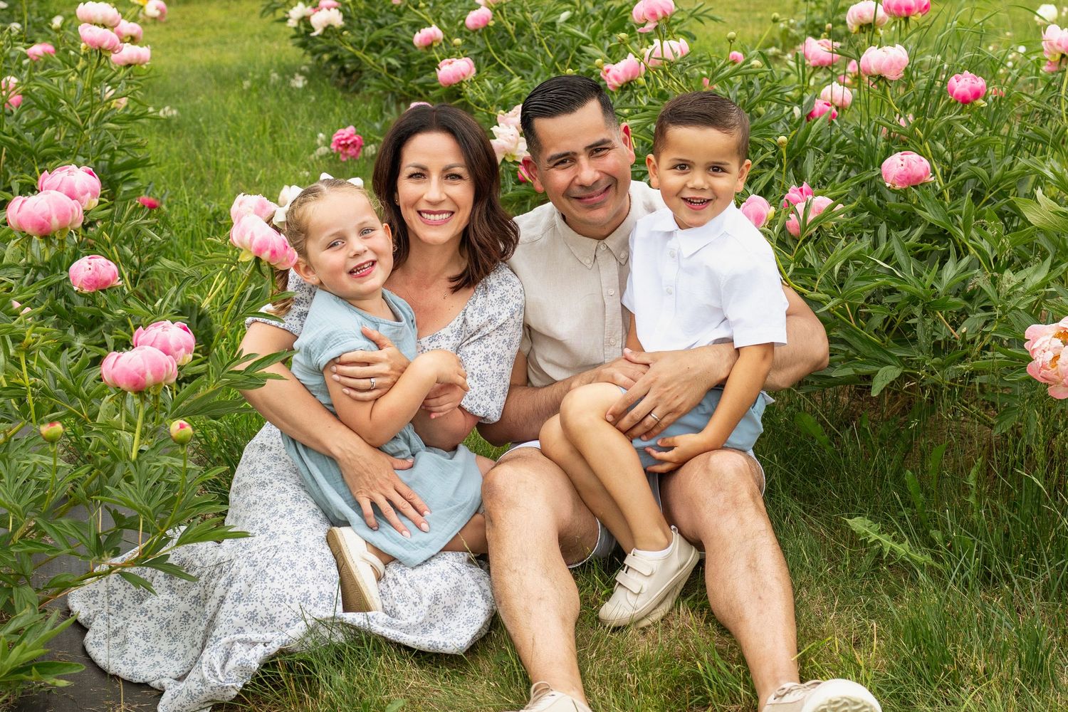 Happy family of four sitting together in a lush garden surrounded by blooming pink peonies.