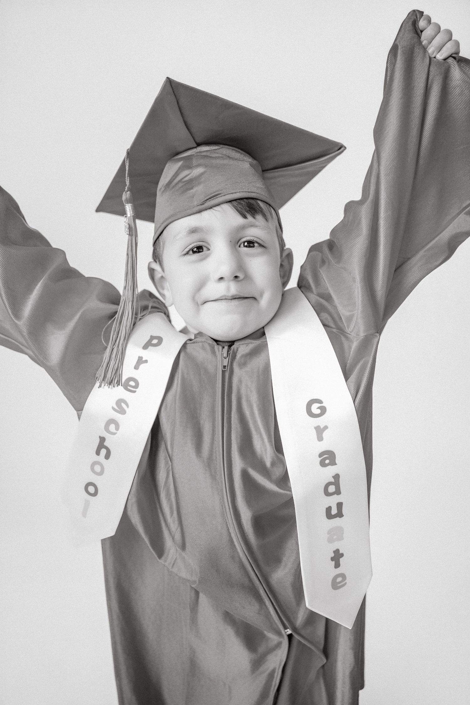 Excited child in preschool graduation cap and gown with arms raised in black and white photo.