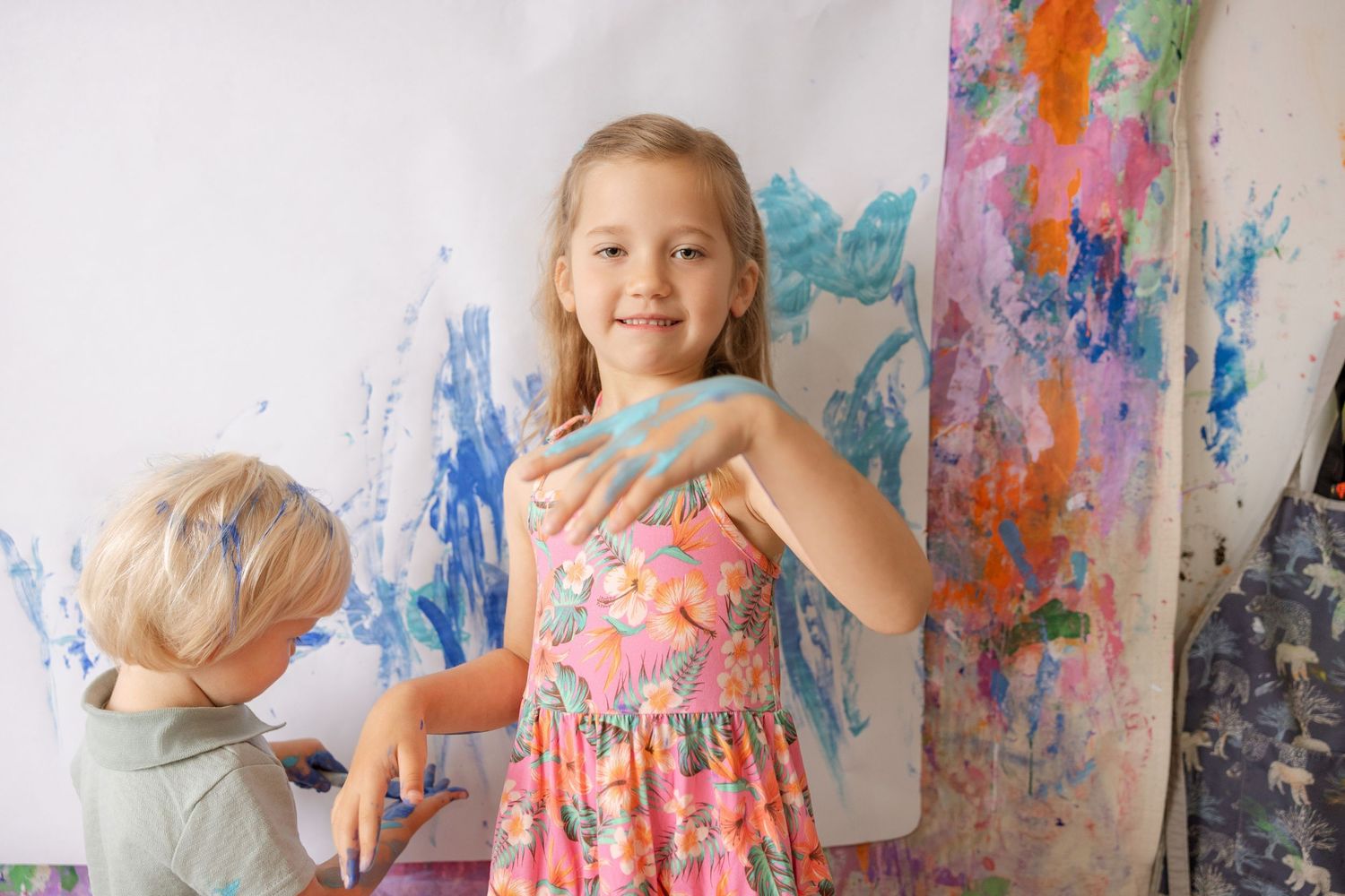 Two children finger painting on a large white canvas, smiling girl in floral dress shows blue paint on her hands.