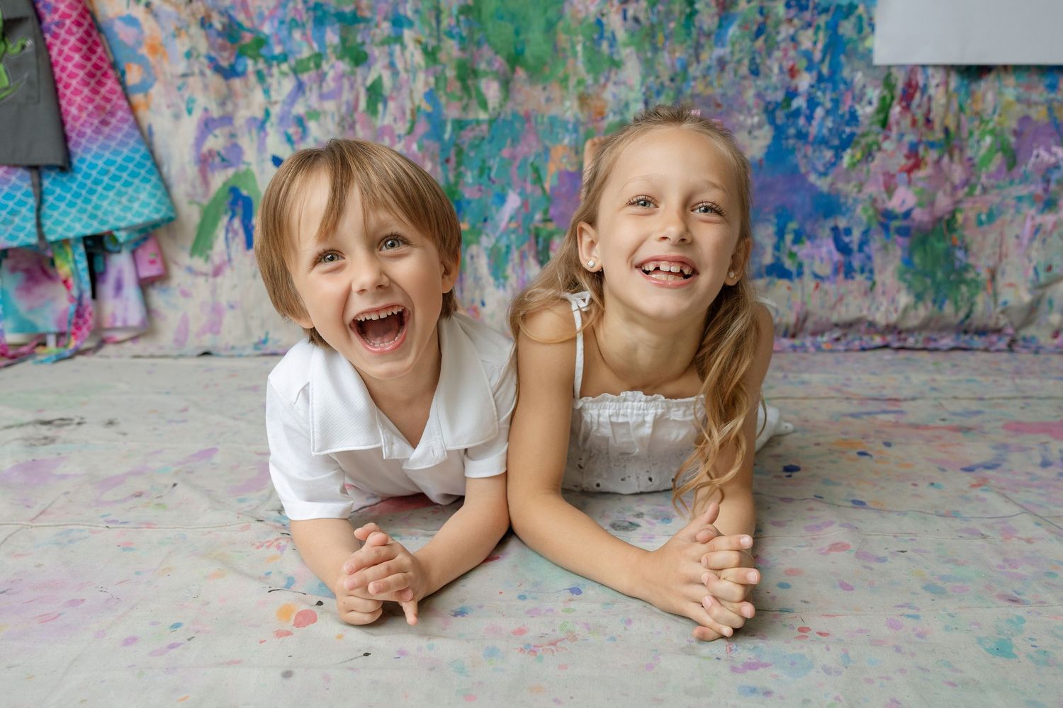 Two happy children laughing and lying on a colorful paint-splattered floor in a vibrant art studio.