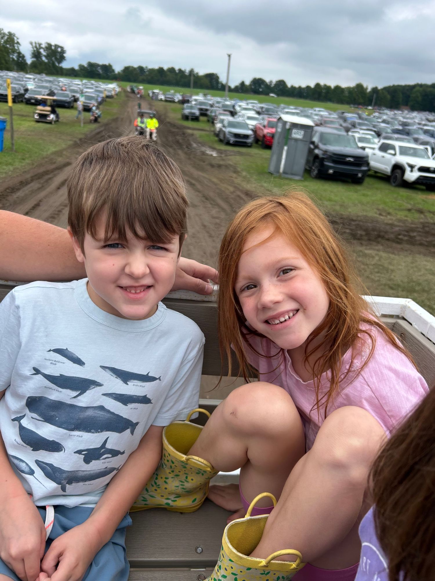 Two smiling children, a boy in a whale shirt and a red-haired girl, sit together at an outdoor muddy event parking area.