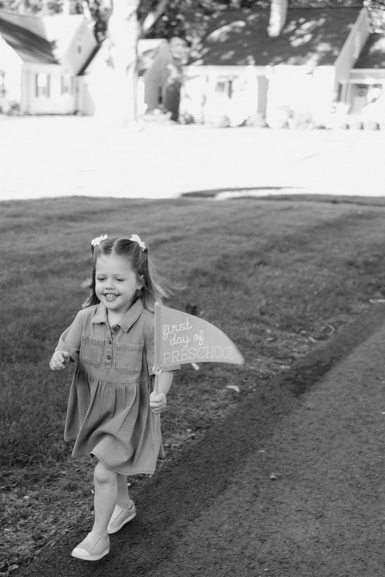 Black and white photo of a smiling young girl in a dress holding a first day of school sign while walking outdoors.