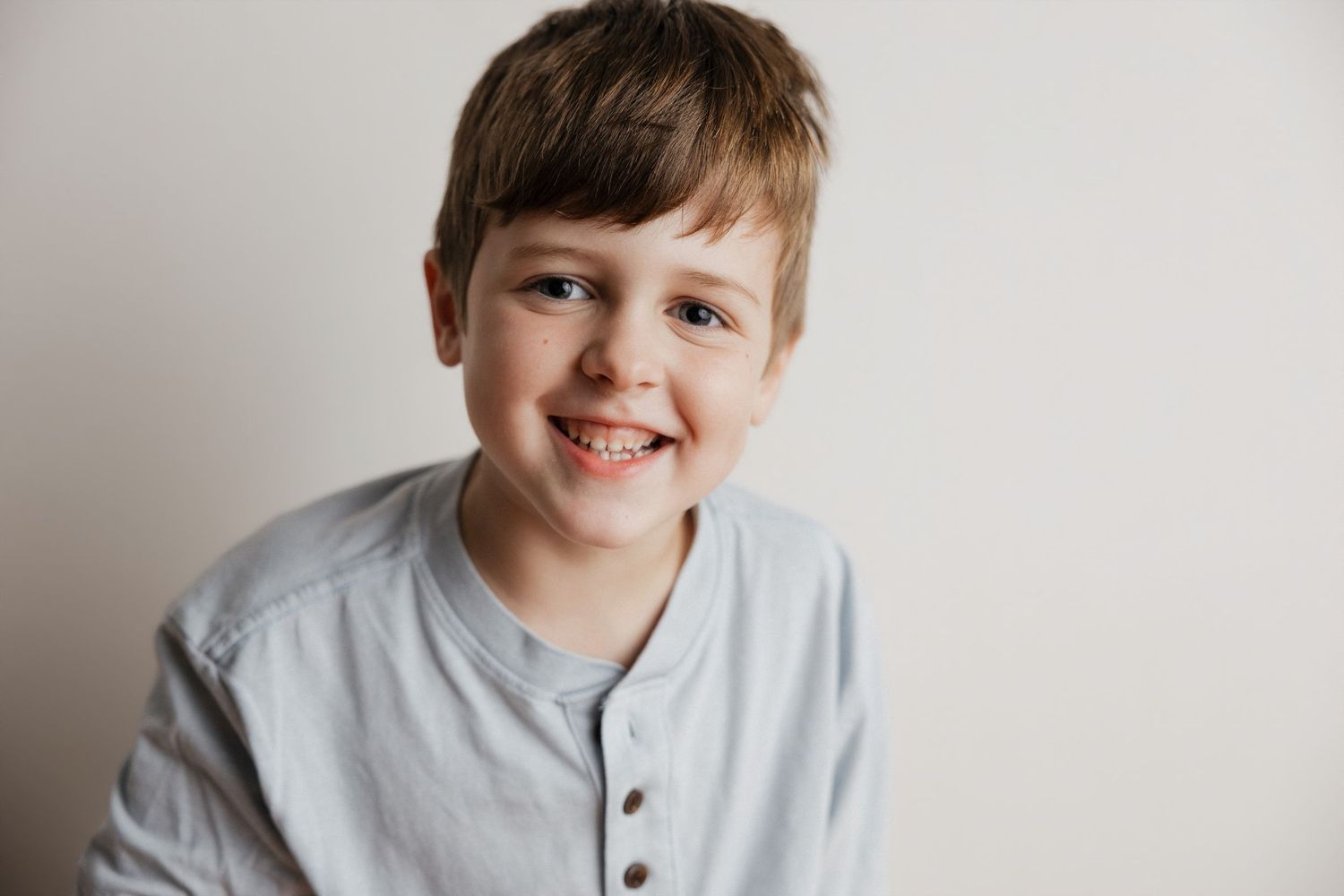 Smiling young boy with brown hair wearing a light gray henley shirt against a white background.