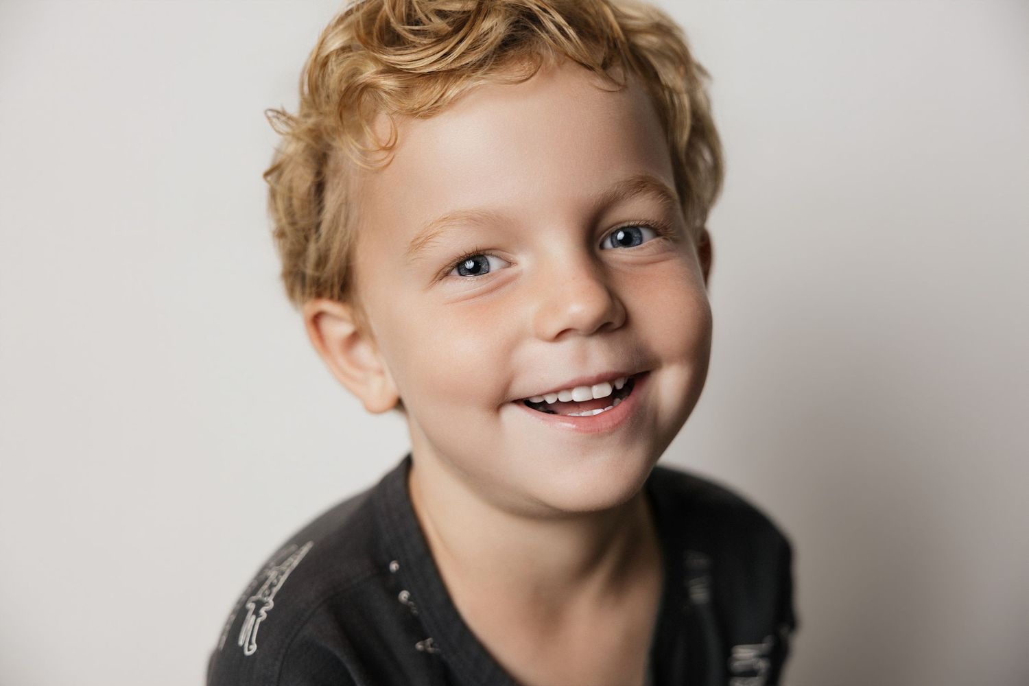 Smiling young boy with curly blond hair wearing a dark graphic tee, studio portrait with white background.