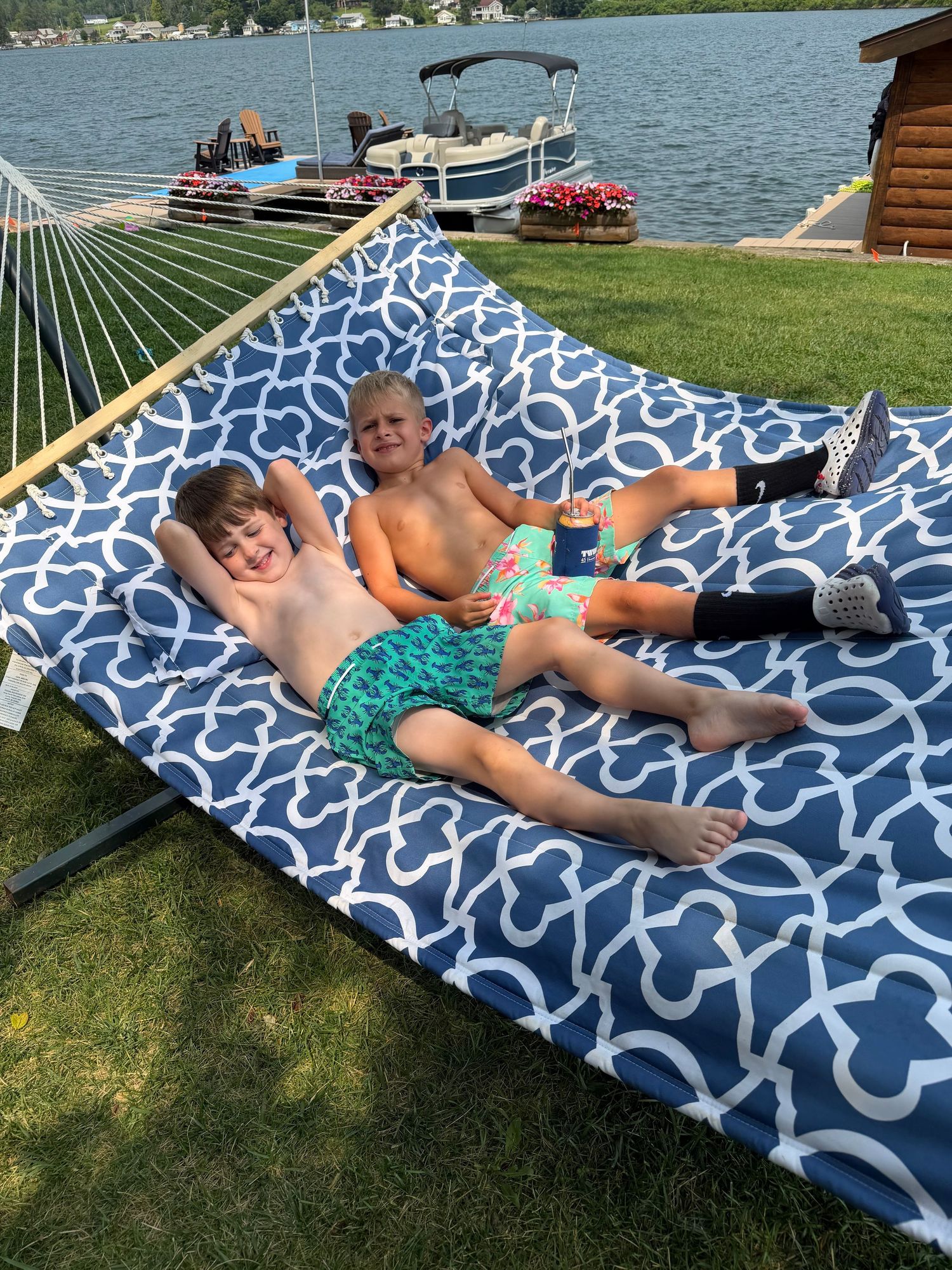 Two children relaxing on a blue patterned hammock by a lakeside on a sunny summer day.