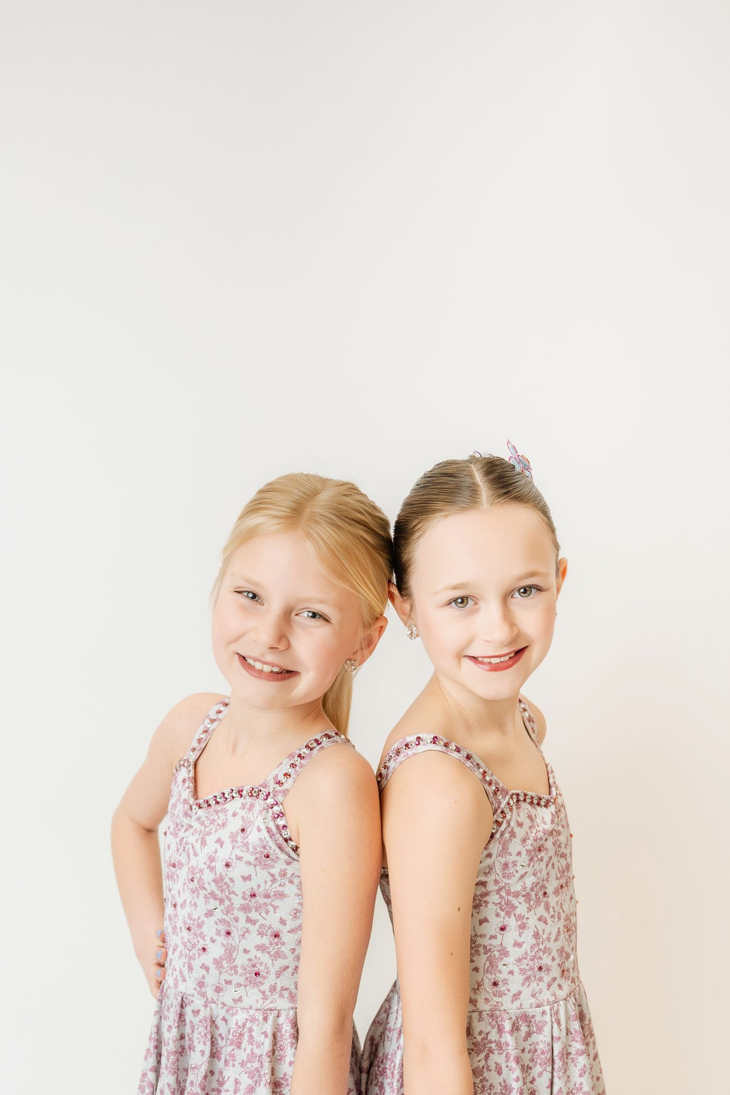 Two smiling girls in matching floral dance dresses standing back-to-back against white background.