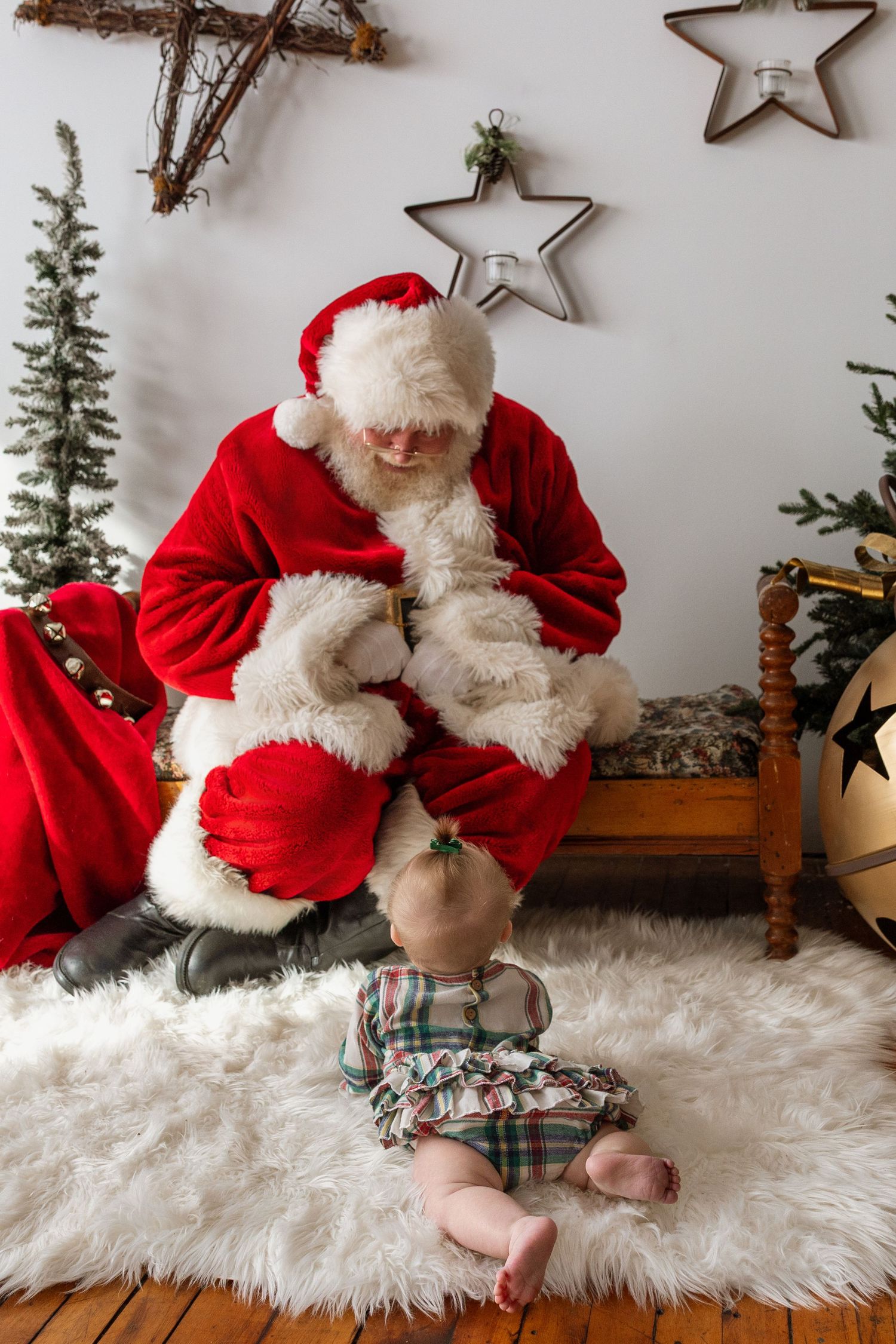 Baby in plaid dress sits on fur rug before Santa Claus in cozy Christmas decorated studio scene.