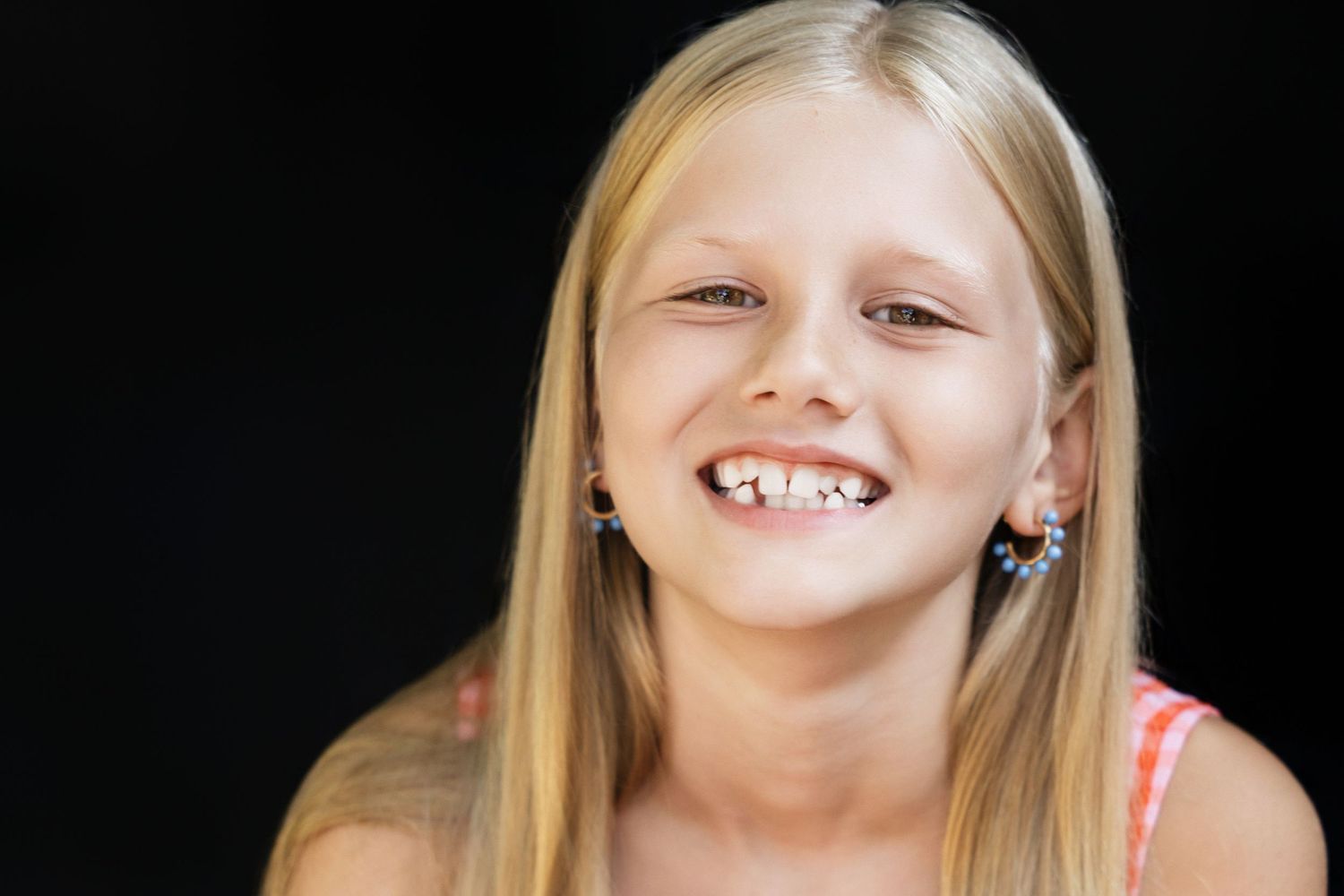 Smiling blonde girl with earrings laughing against black background, showing missing teeth in a cheerful portrait.
