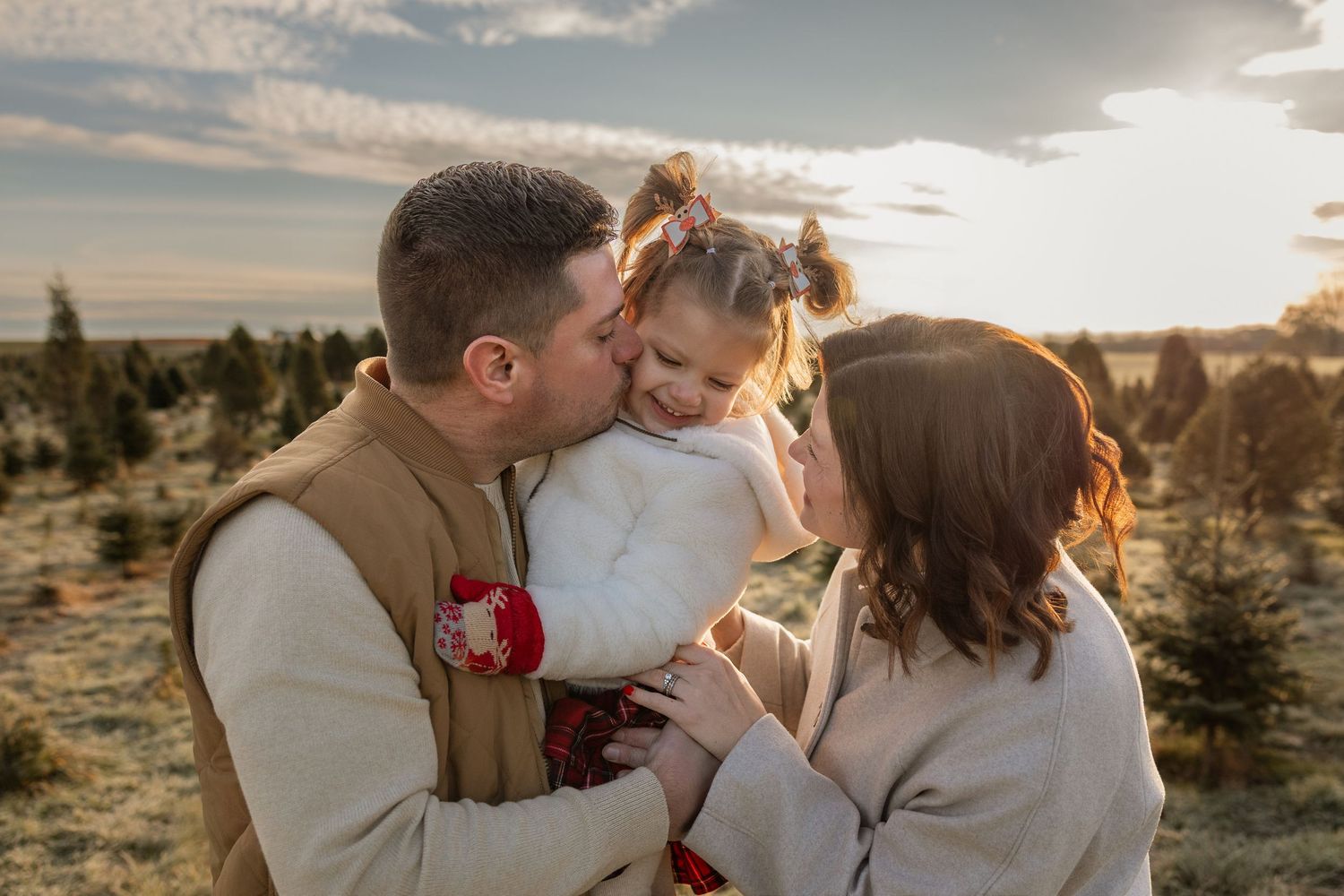 A family of three shares a tender moment at sunset in a Christmas tree farm, parents kissing their young daughter.