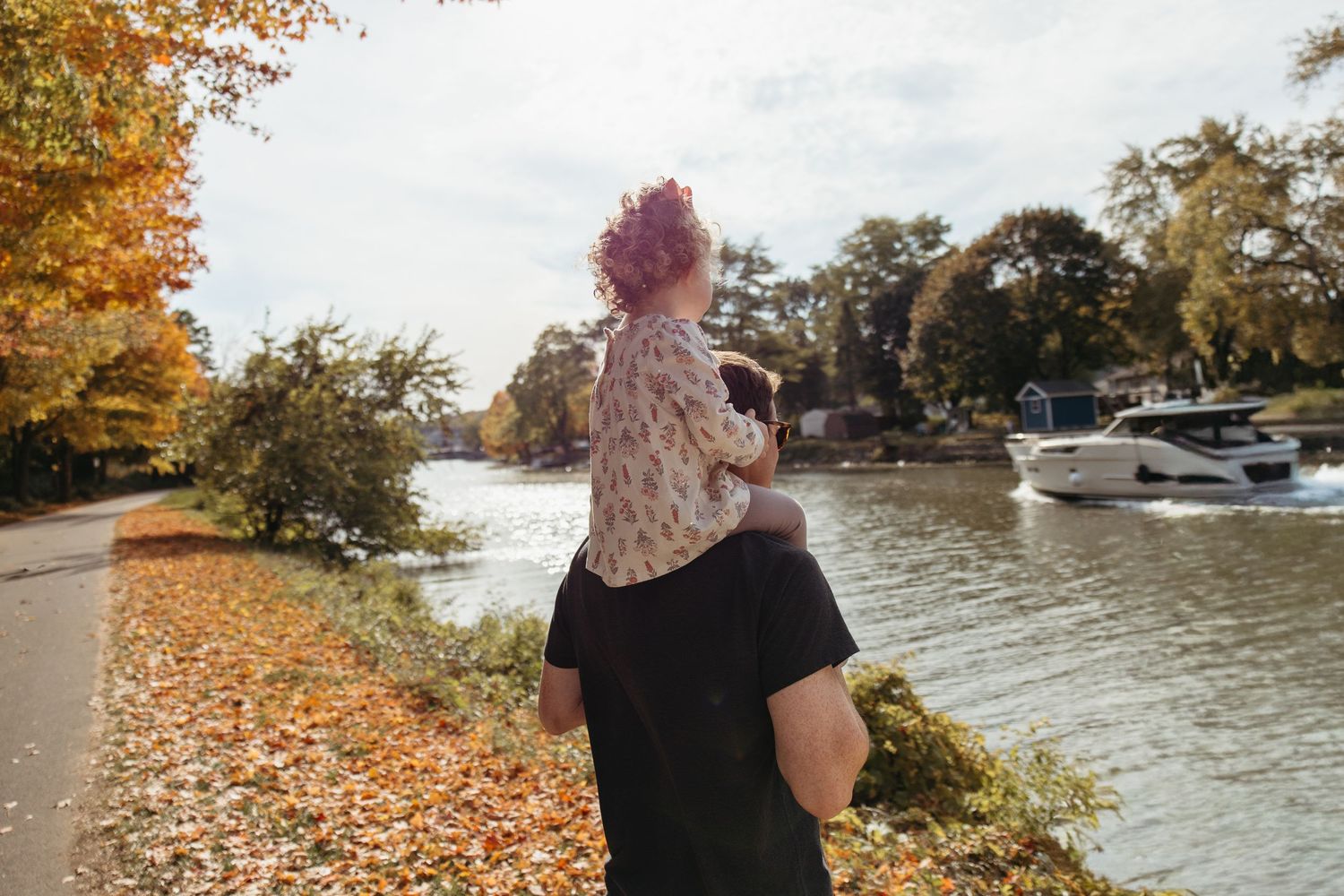 Father carrying young child on shoulders along a scenic autumn riverside path with colorful fall foliage.