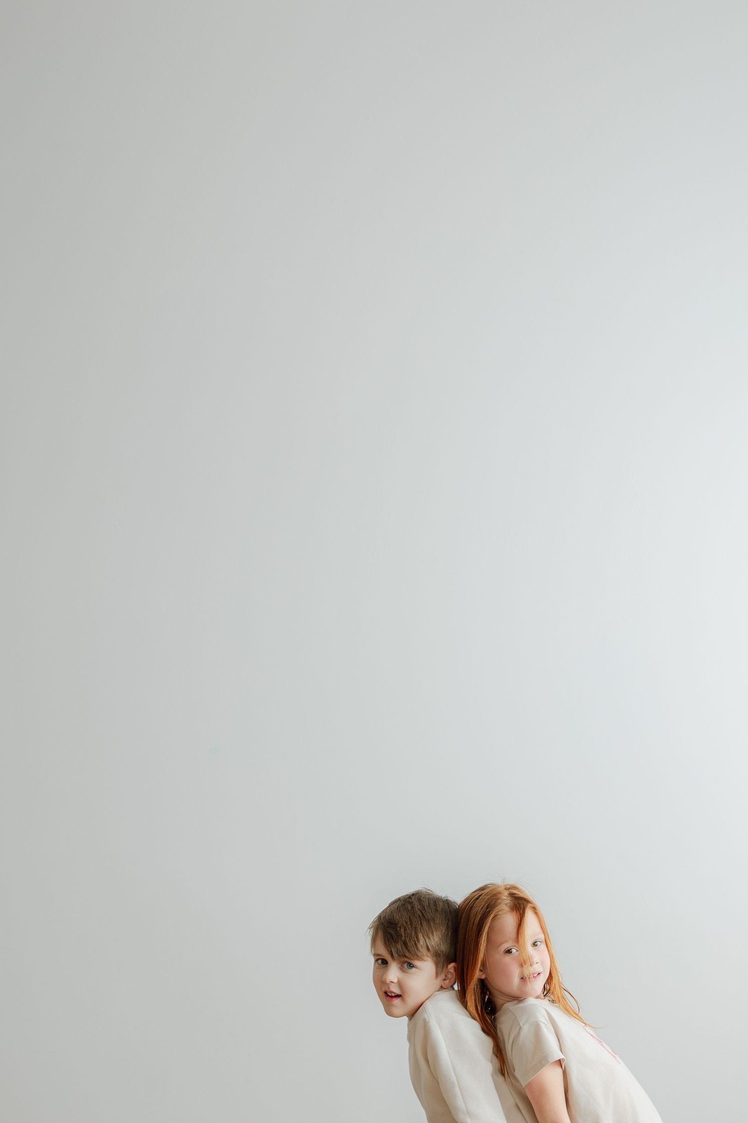 Two young children in white outfits pose back-to-back against a light gray background.