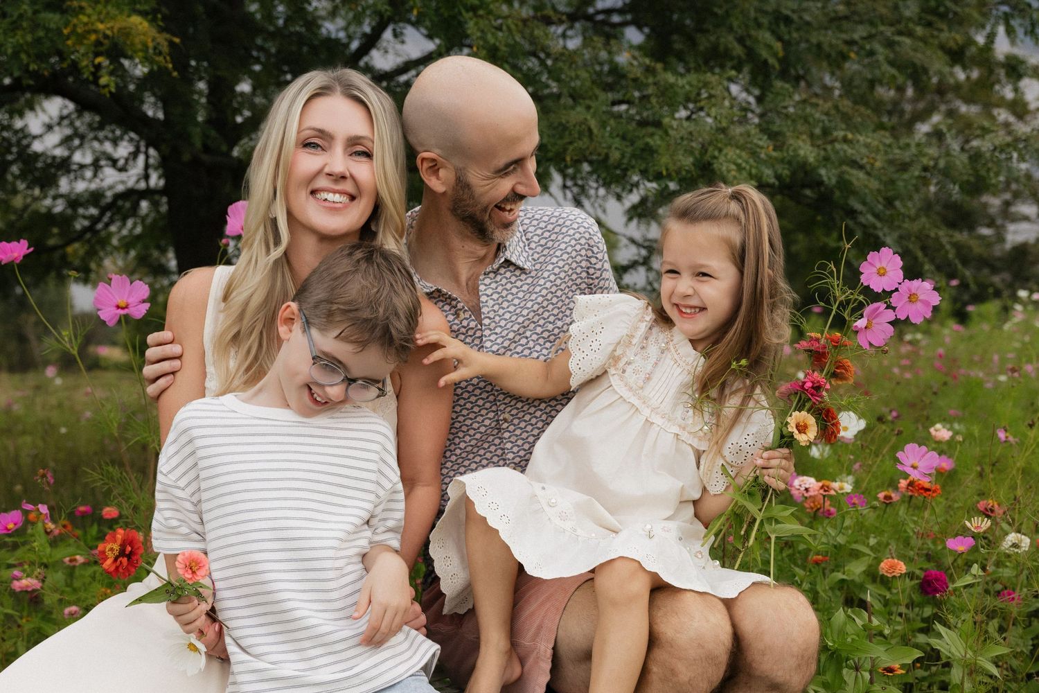 Family of four smiling together in a flower garden, with pink and white blooms surrounding them on a sunny day.