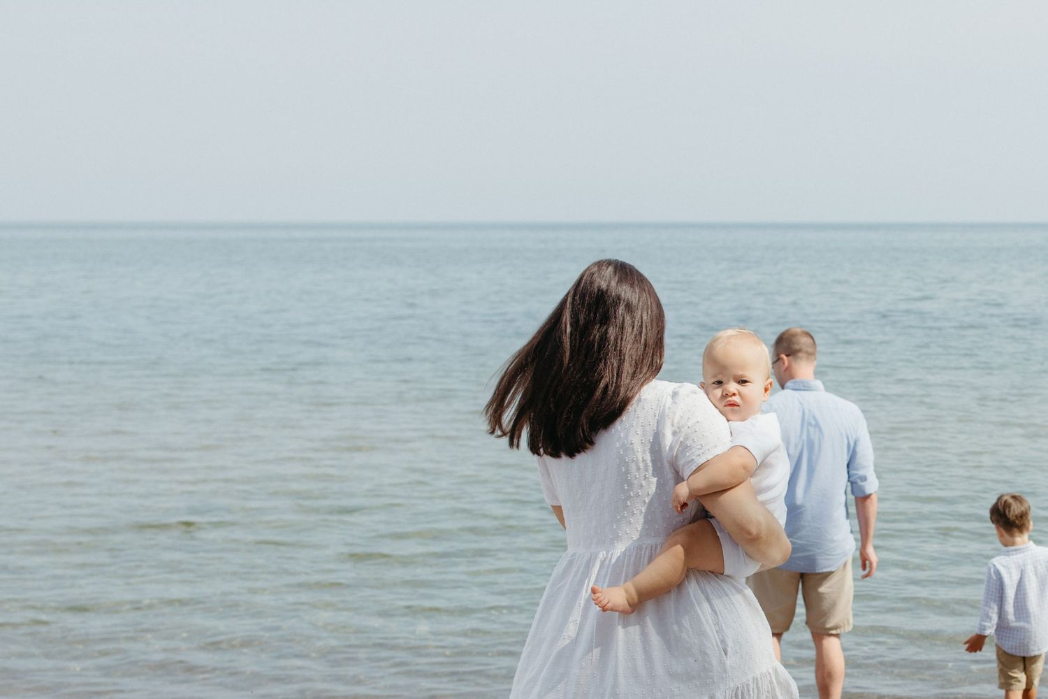 Family walking on beach, mother in white dress holding baby, father and child strolling toward calm ocean on overcast day.