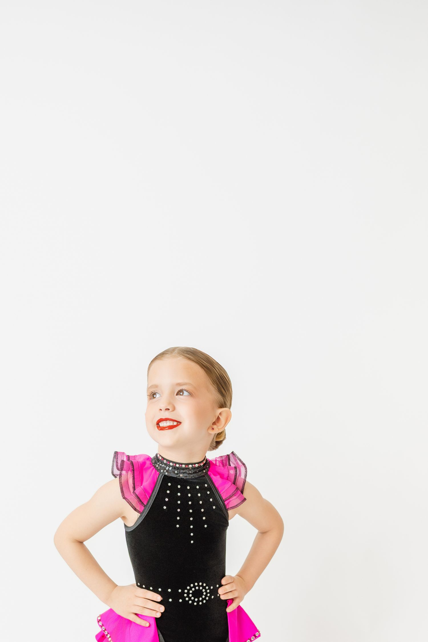 Young dancer in black and pink rhinestone costume poses with hands on hips looking upward.