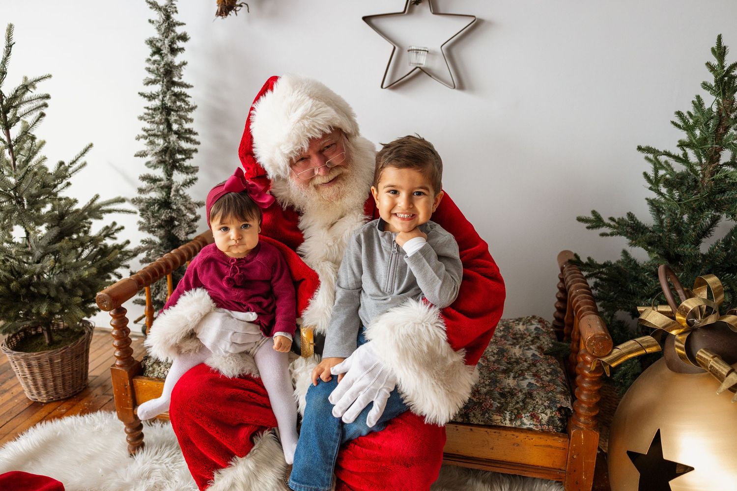 Santa Claus sits on bench holding two smiling children in bright Christmas studio with gold bell.