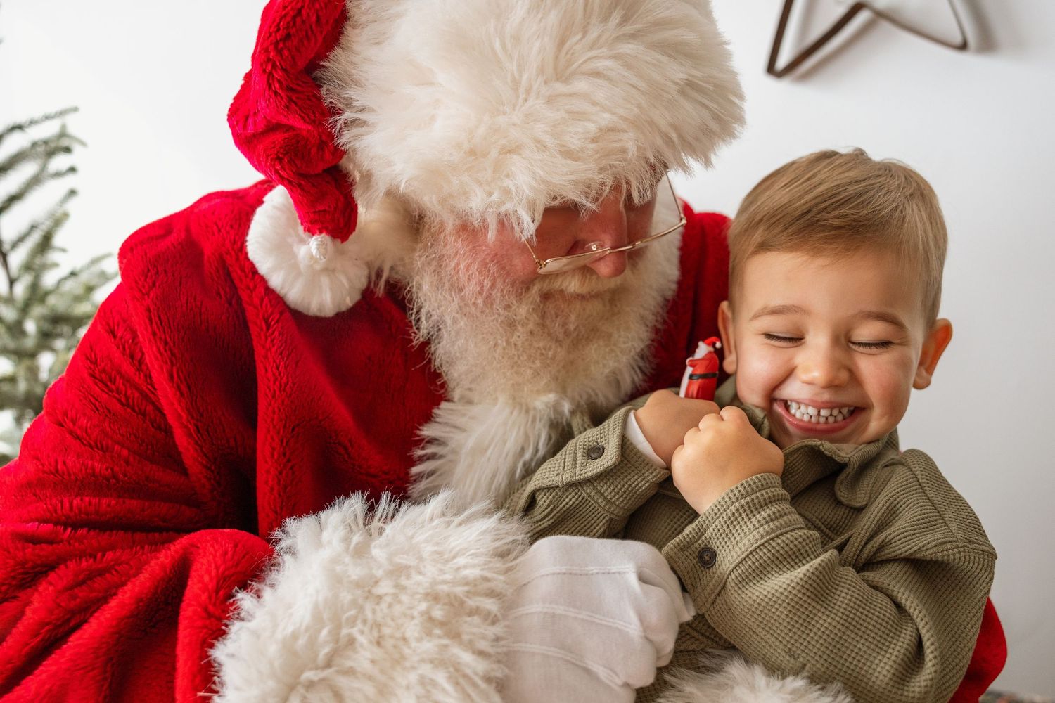 A delighted toddler in a green jacket laughs while sitting with Santa Claus during a Christmas photo session.