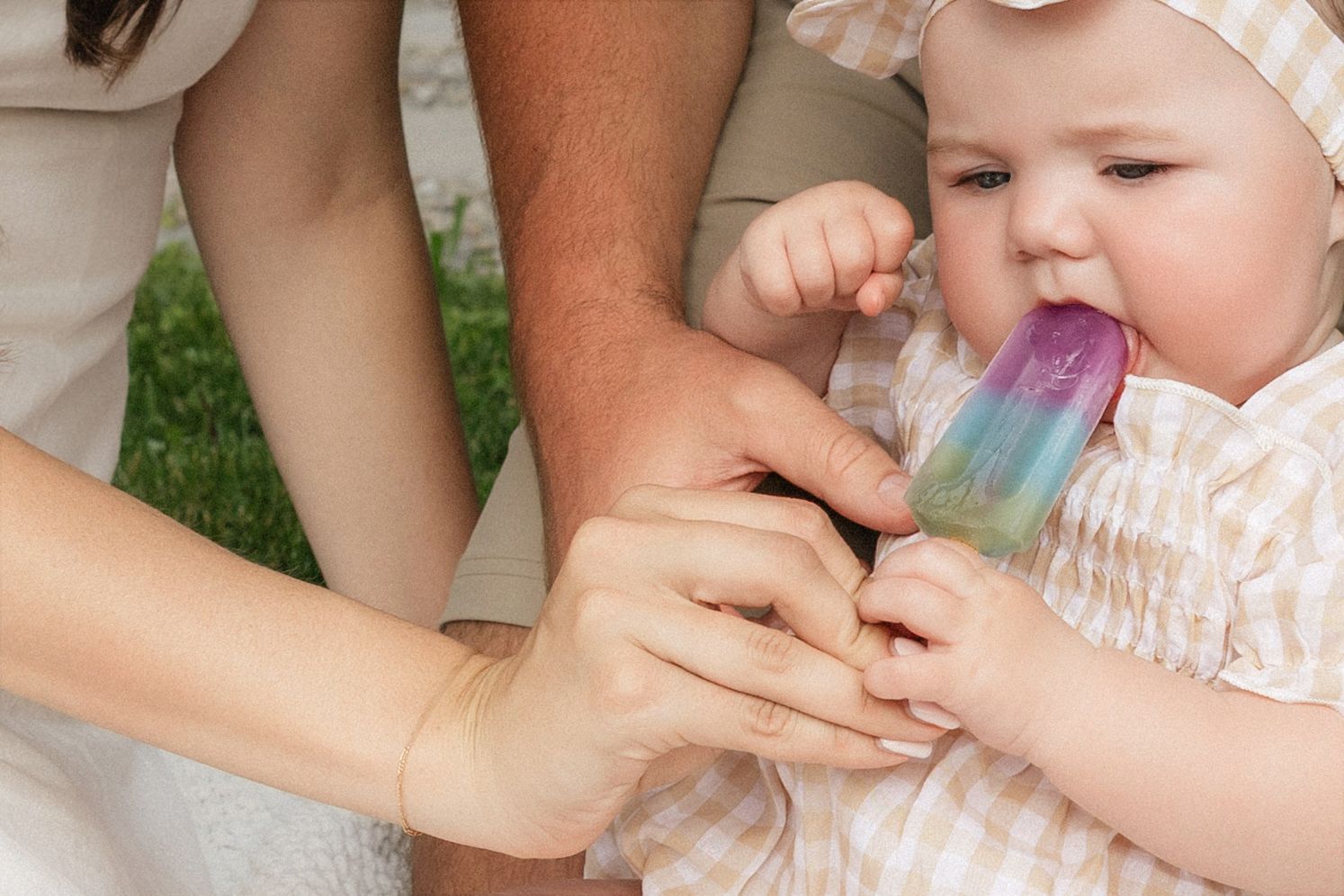 Baby tasting a colorful rainbow popsicle held by parents outdoors in summer.