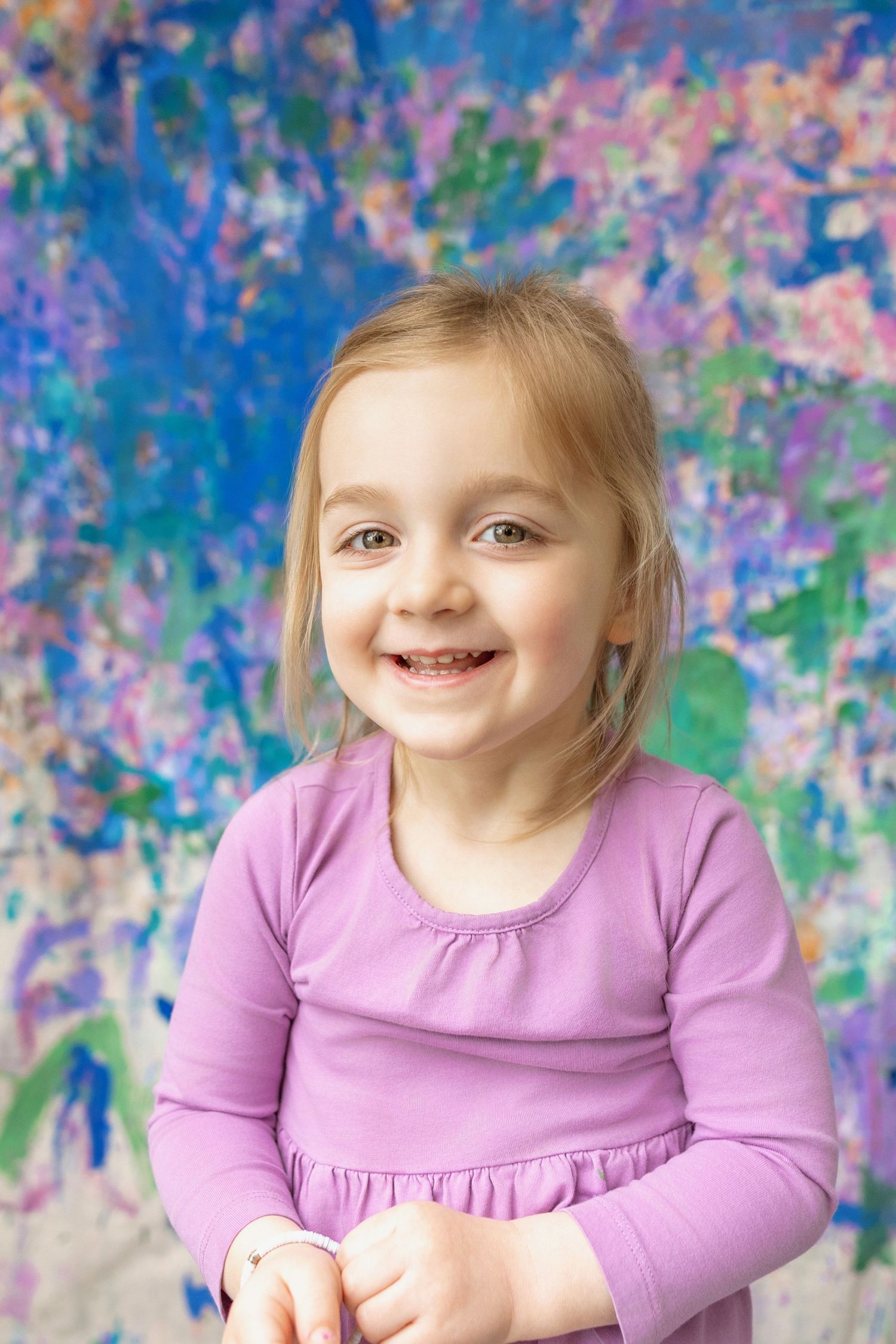 A cheerful young girl in a purple dress smiles broadly against a colorful abstract background.