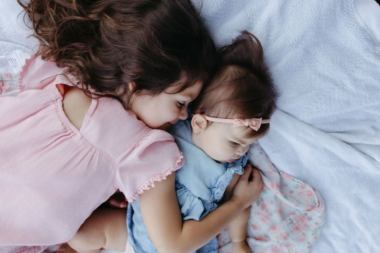 Two young sisters sleeping peacefully together on a white blanket, one in pink and one in blue.