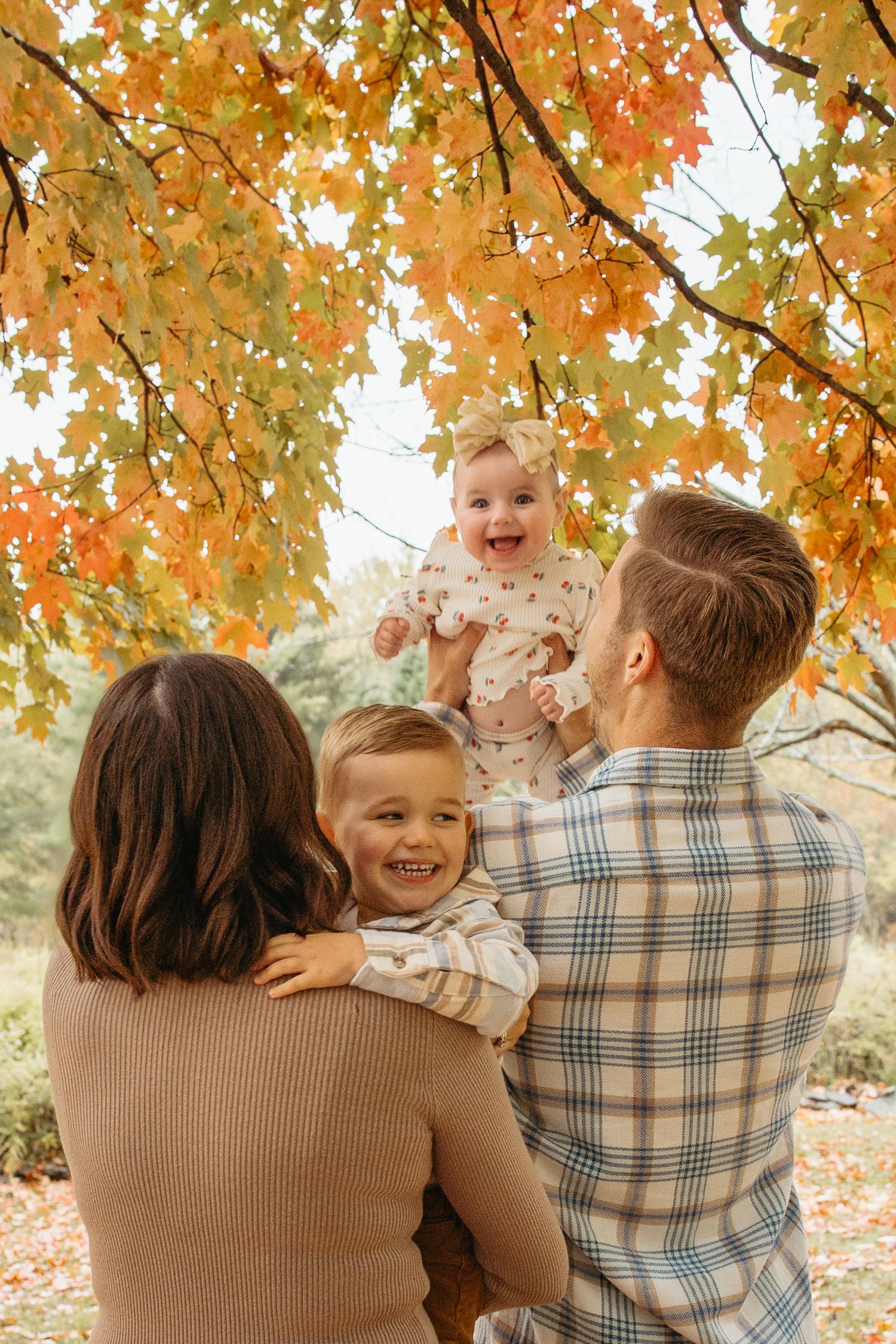 Family of four enjoying autumn park with baby held up under vibrant orange maple leaves.