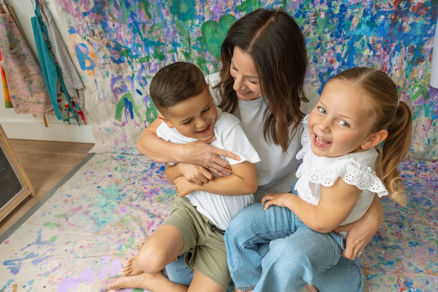 A mother sits on a colorful painted floor hugging her two young children, smiling joyfully.