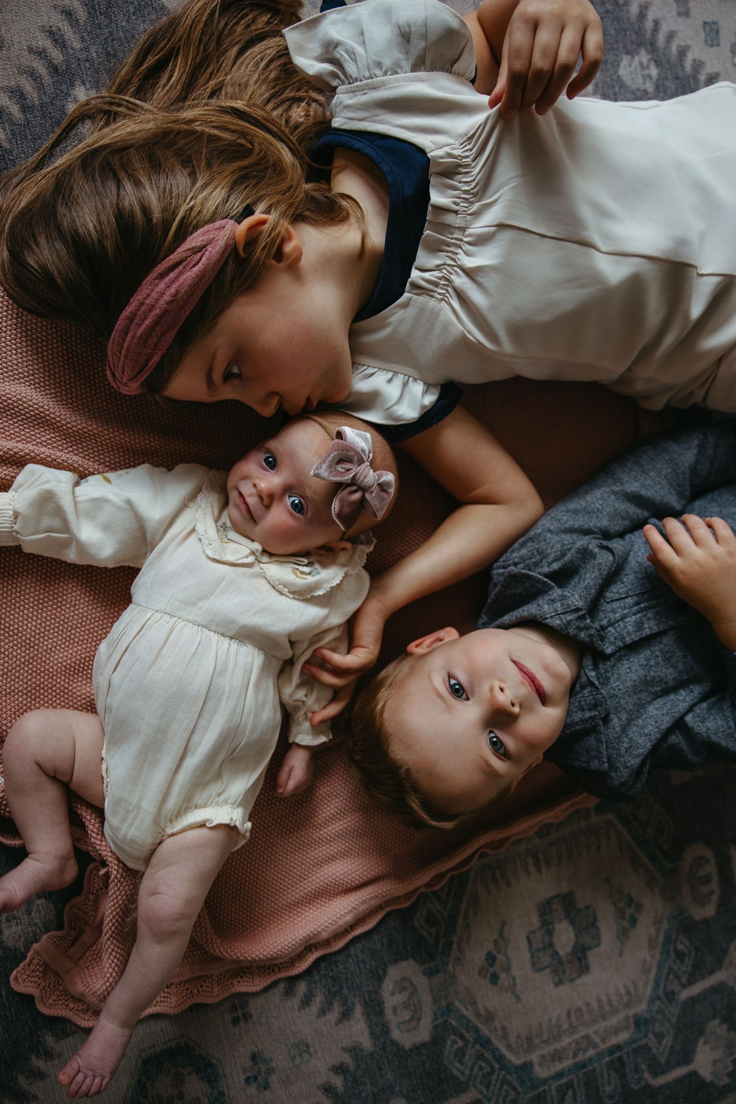 Three siblings lie together on colorful blankets, older girl kissing newborn baby sister tenderly.