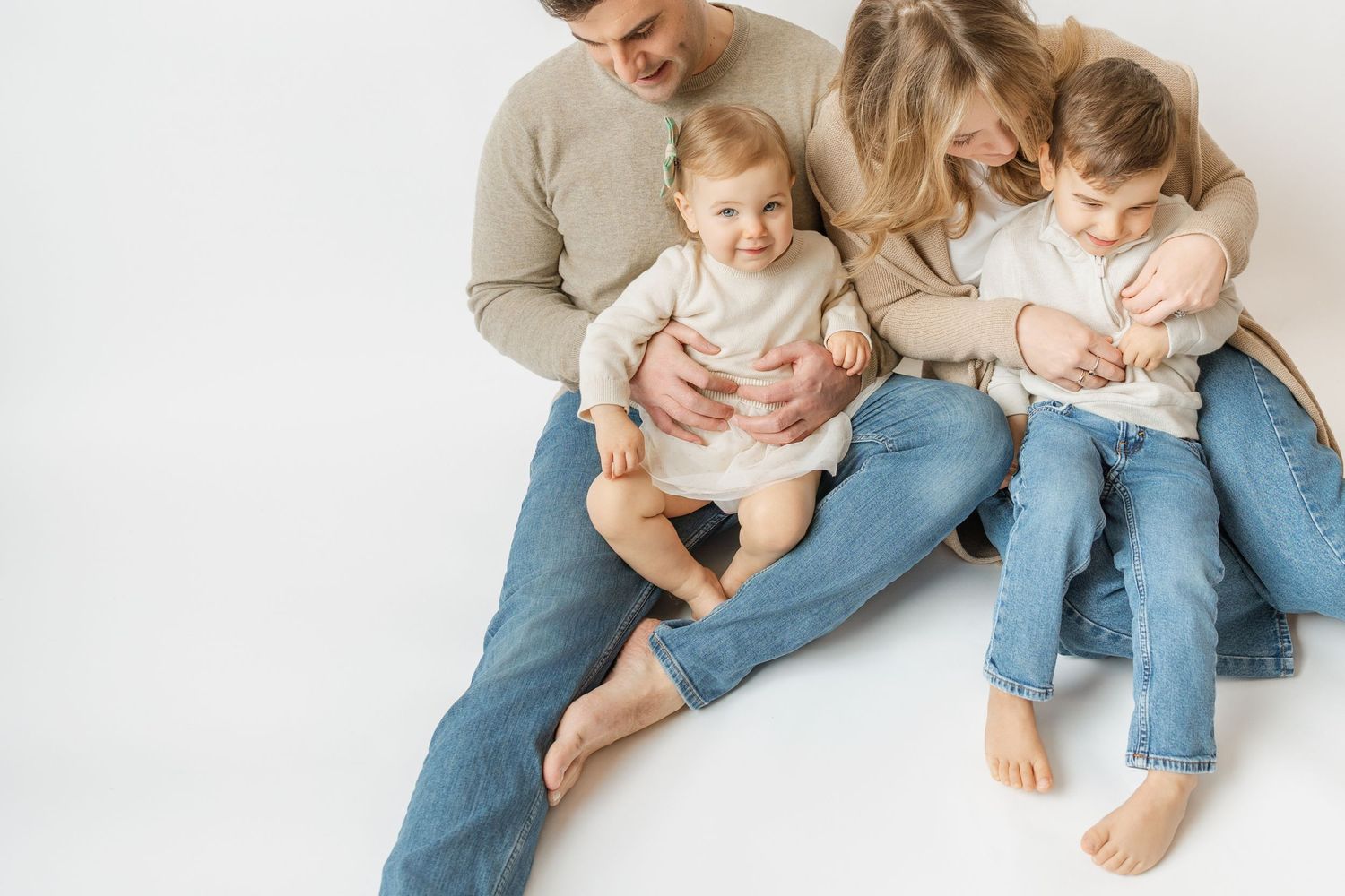 Happy family of four sitting on white floor, parents with two young children in jeans.