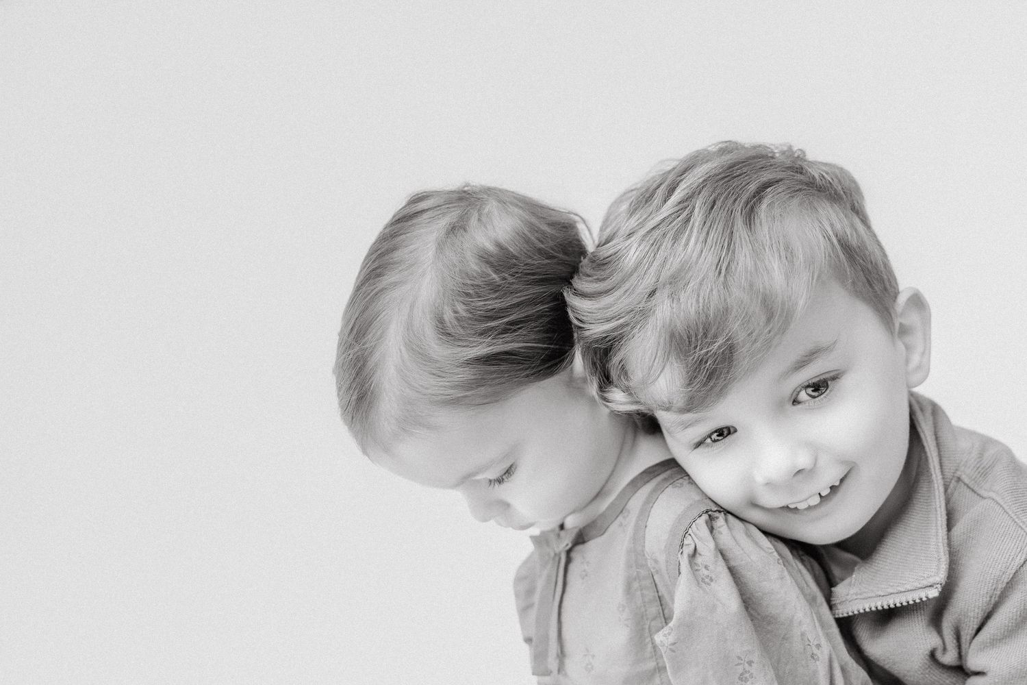Black and white portrait of two young siblings leaning heads together smiling.
