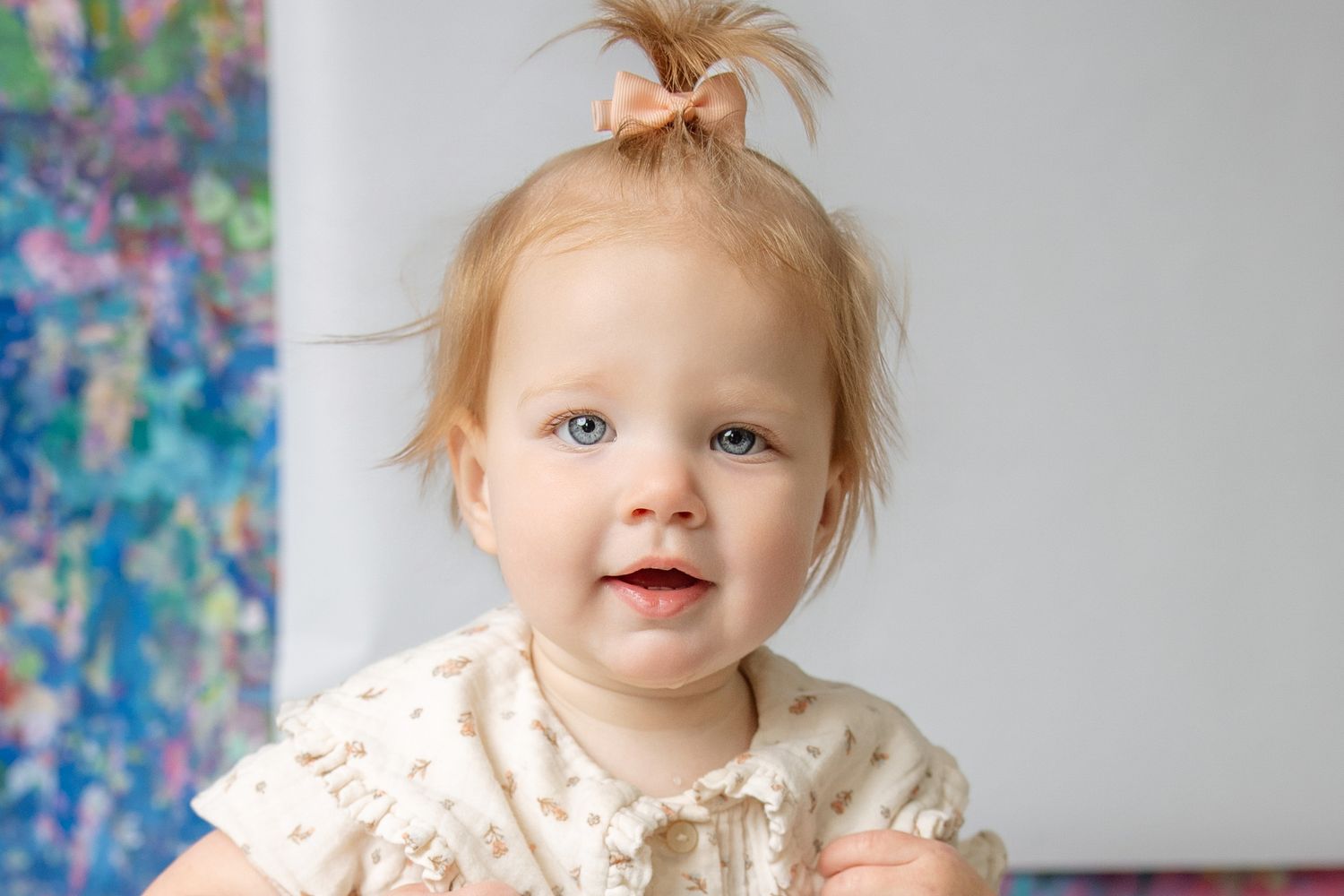 Adorable baby girl with blue eyes and tiny hair bow smiles sweetly in a white dress.