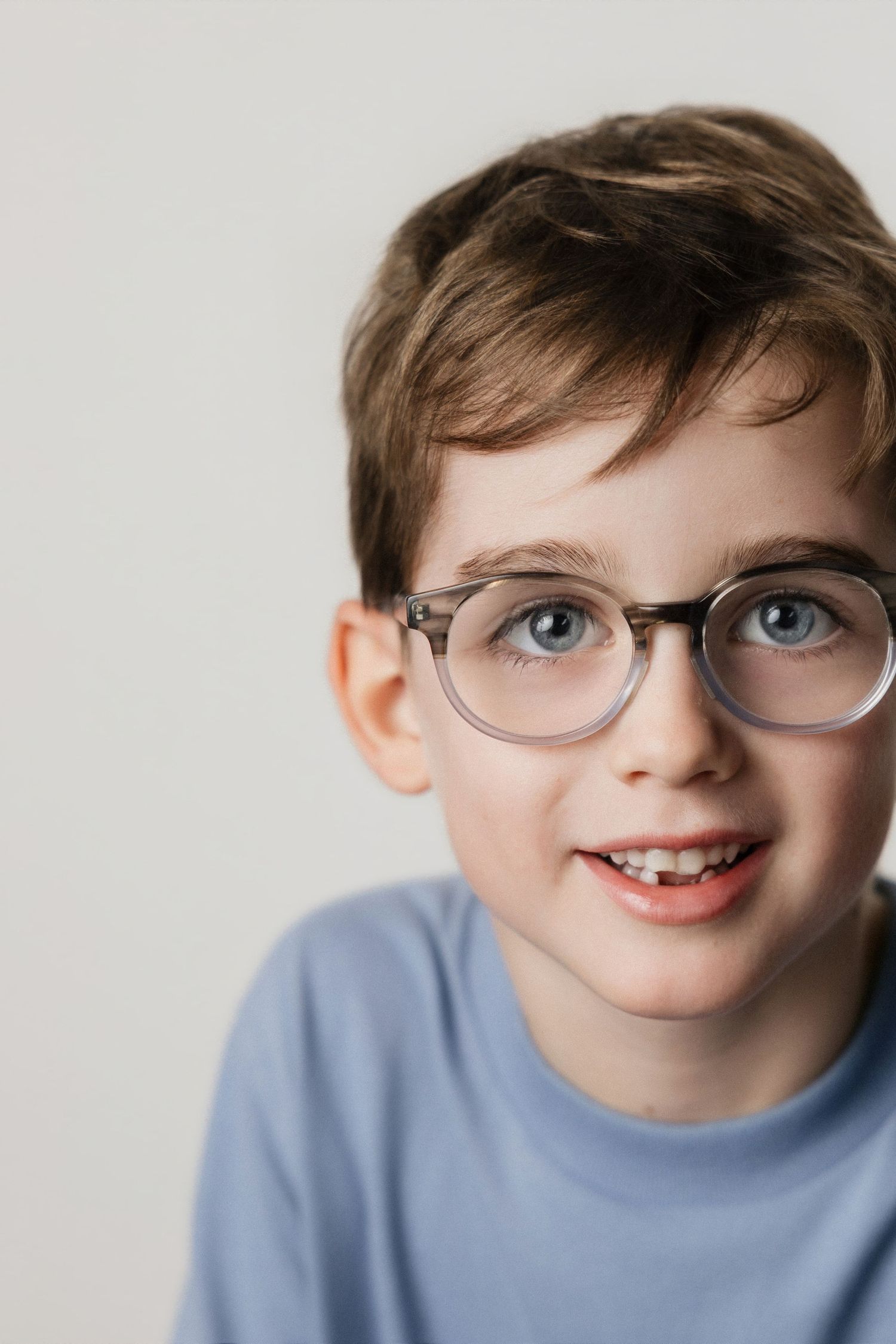 A smiling young boy with blue eyes wearing round gray glasses and a light blue t-shirt against a white background.