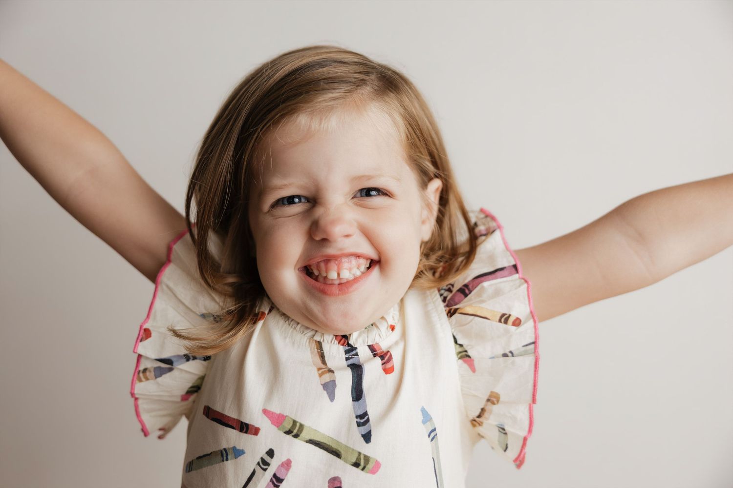 Happy toddler girl in crayon-print dress smiling with arms outstretched against white background.