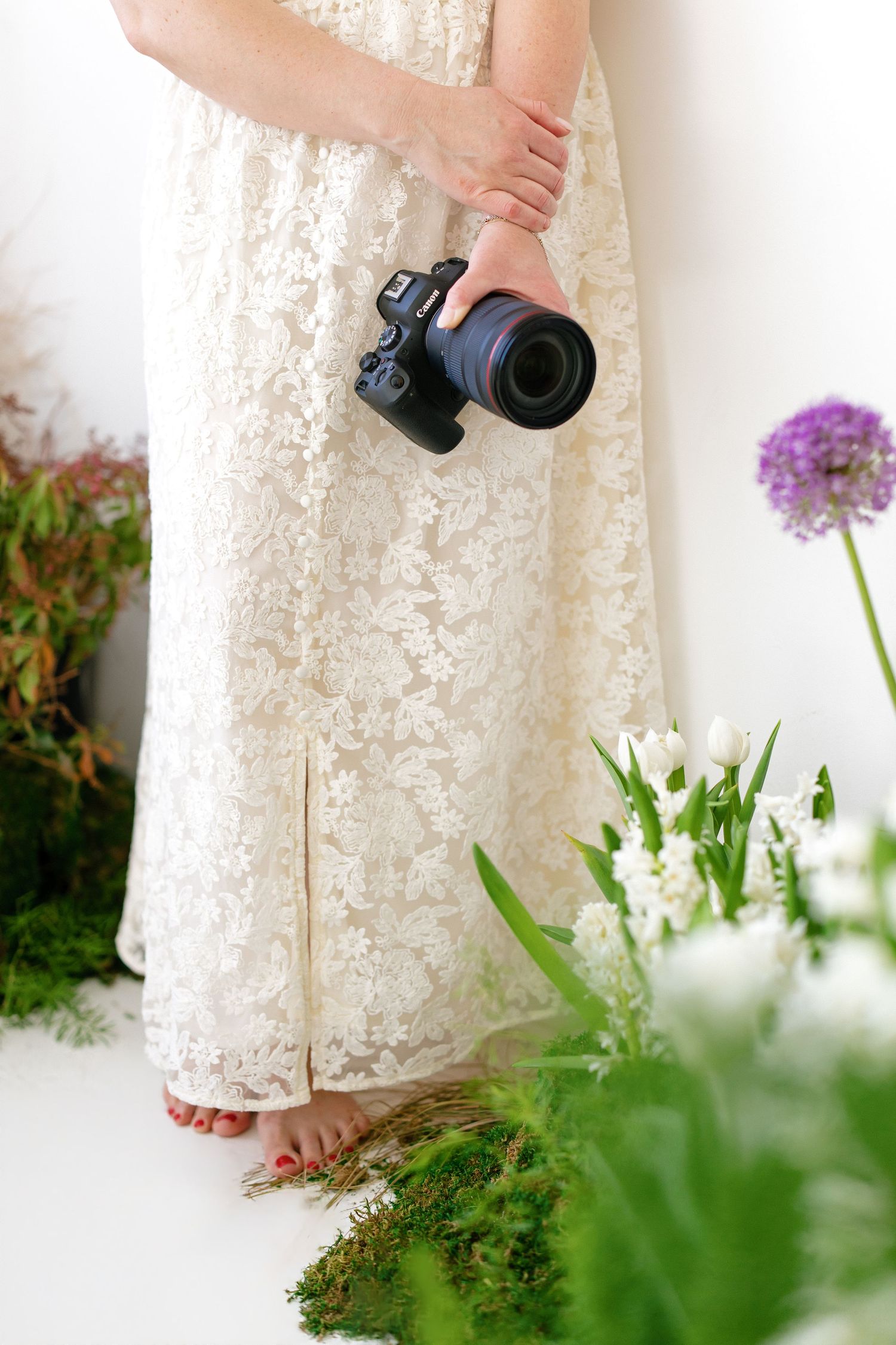 Hilary Mauro in white lace dress holding a DSLR camera surrounded by fresh spring flowers in a bright studio.