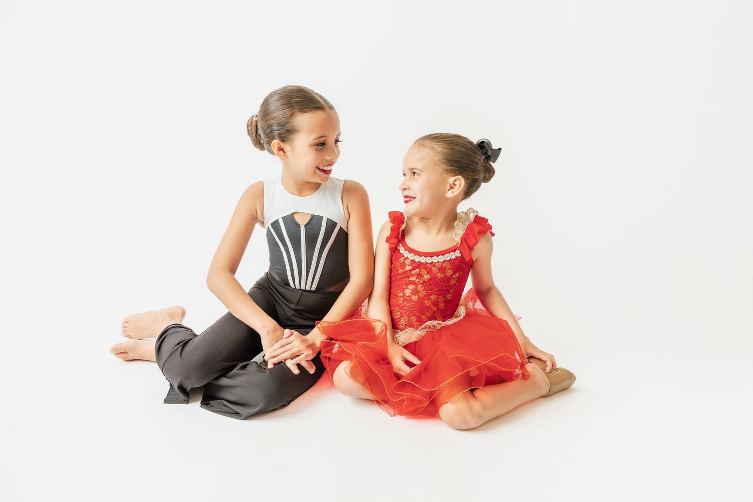 Two young girls in dance costumes sitting together laughing on a white background.