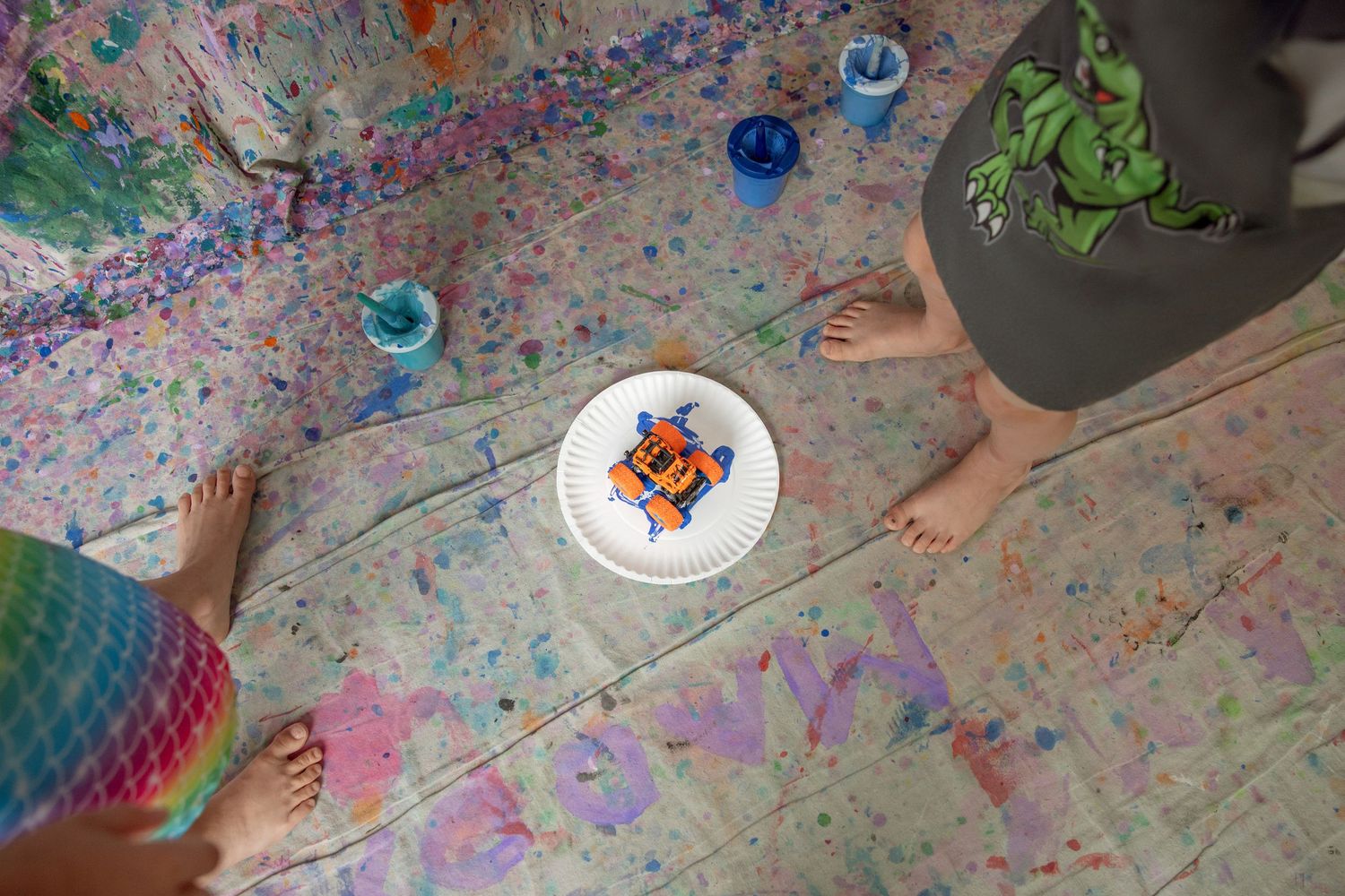 Overhead view of two barefoot children standing on a paint-splattered canvas floor with a decorated paper plate.