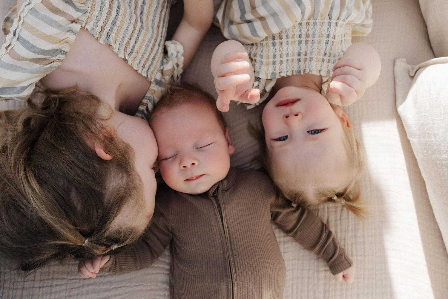 Three young children lying together on a cream blanket, including a sleeping newborn baby between two toddlers.