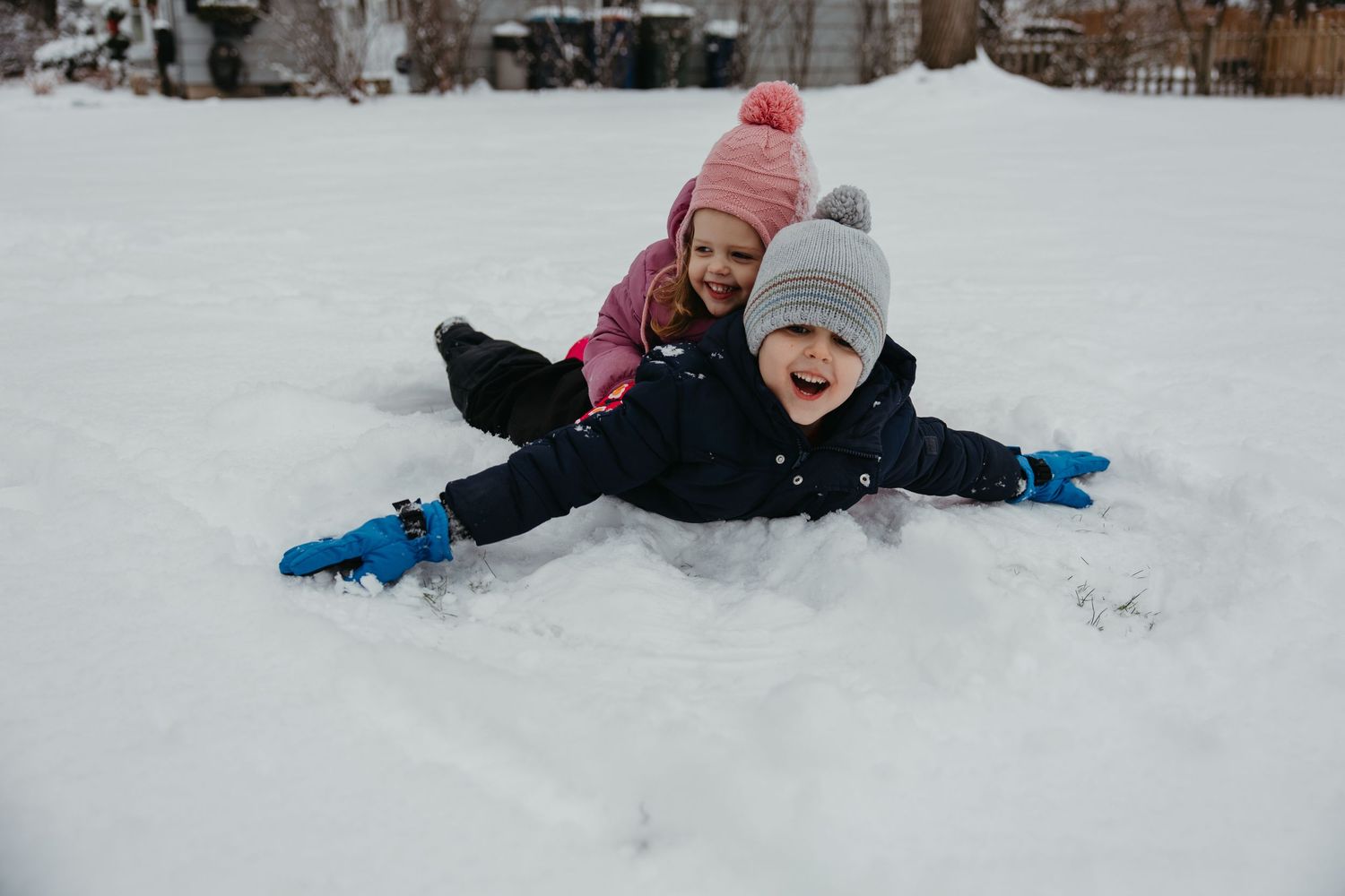 Two children laughing and playing in deep snow during winter wearing colorful hats.