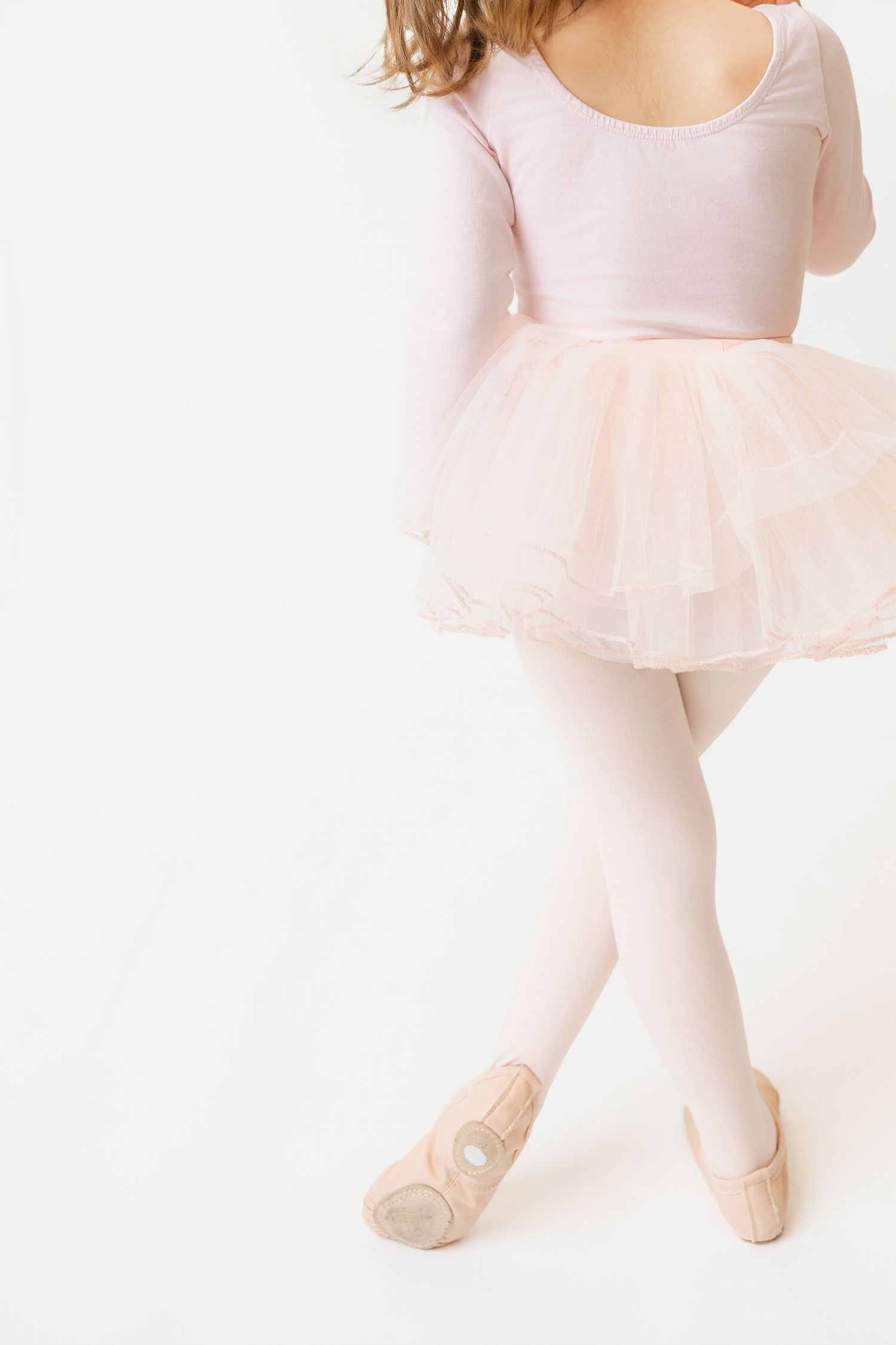 Little ballerina in pink tutu and ballet slippers stands in a classic dance pose on white background.