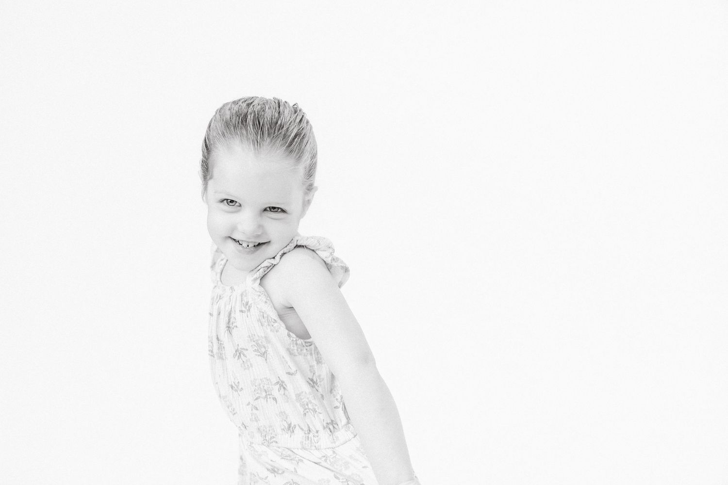 Black and white portrait of a smiling young girl in a floral dress posing against a white background.