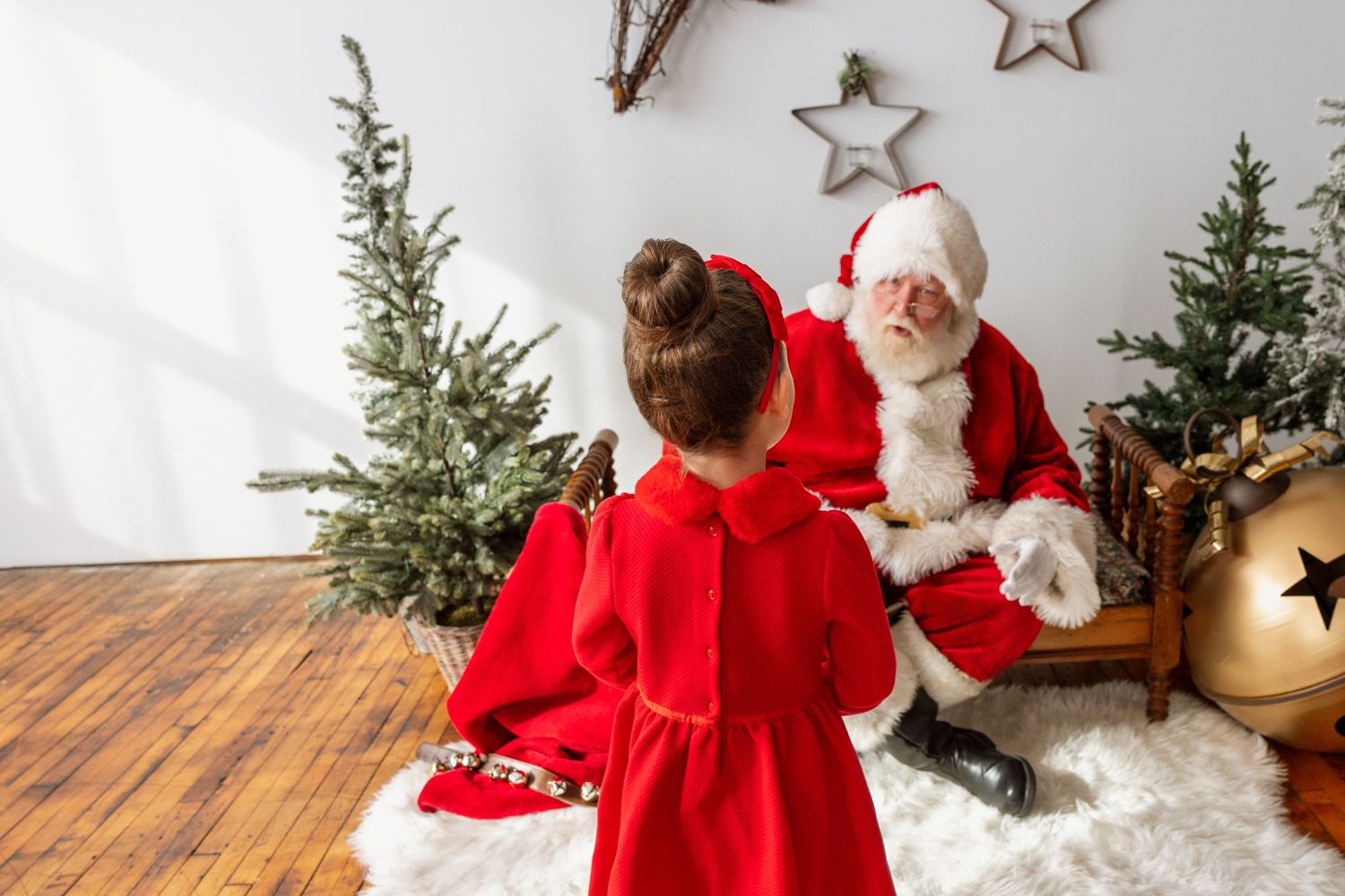 A young girl in a red dress approaches Santa Claus seated near decorated Christmas trees on a fur rug.